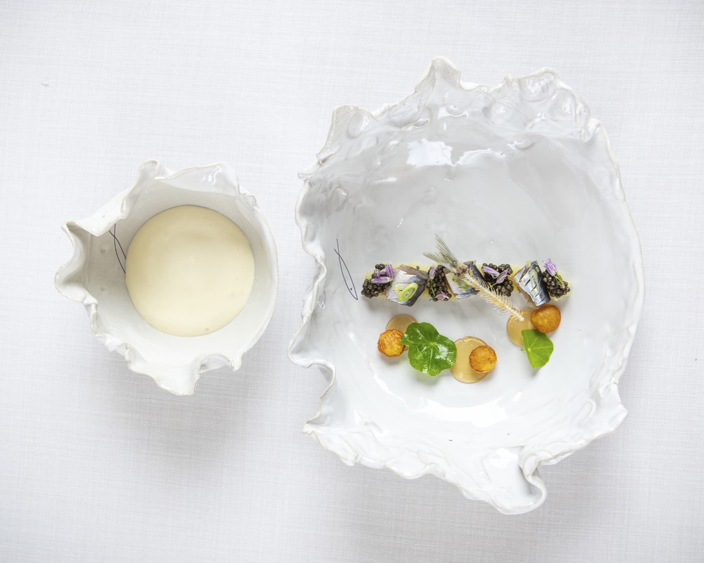 Two oyster-shell-shaped white porcelain bowls — one holding a smooth velouté, the other a row of marinated sardines on caviar with nasturtium leaves and small fried discs.