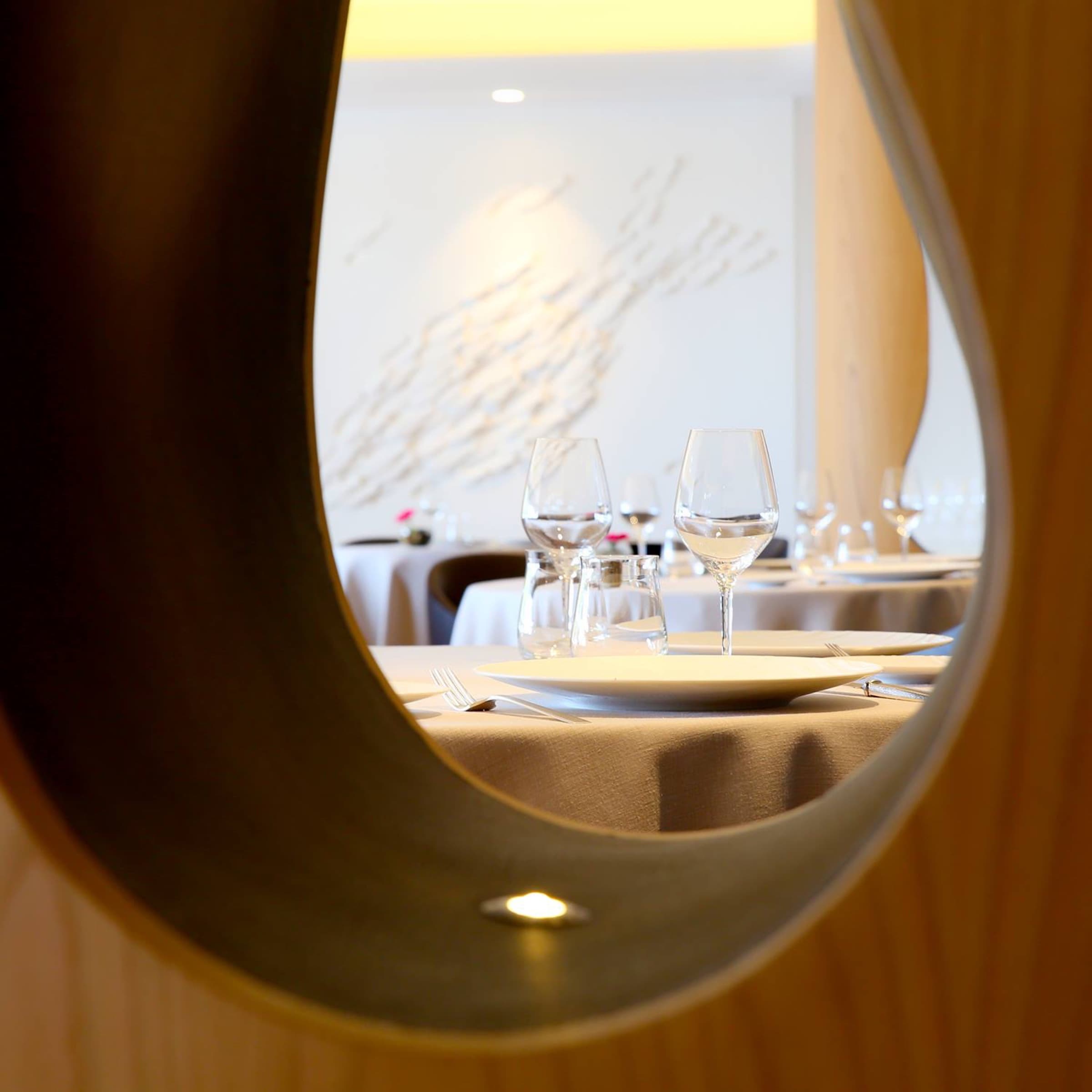 The dining room glimpsed through the curve of one of the room's sculpted oak columns — set tables in white linen, stemware, a shoal-pattern wall sculpture catching warm light.