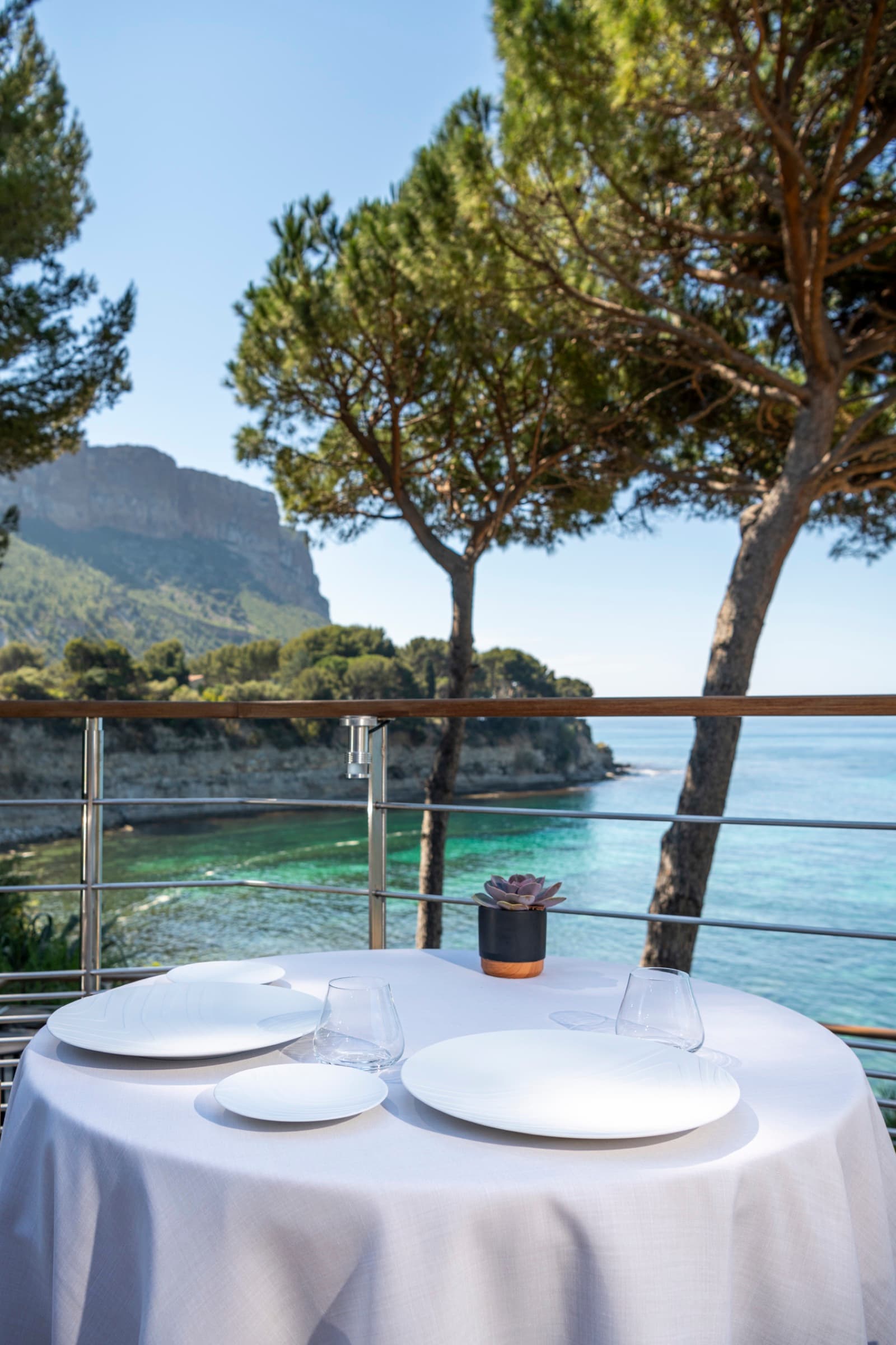 A round table for two laid with three white plates and stemware, a small succulent in a stone pot, the railing of the cliff terrace, the cliffs of Cap Canaille and a stretch of the Mediterranean.