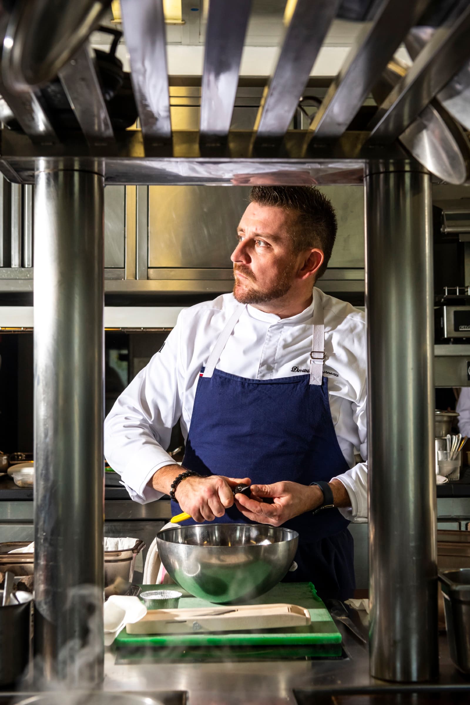 Chef Dimitri Droisneau in his kitchen, blue apron with his name embroidered, looking off-camera, working with a small bowl on a stainless-steel pass.