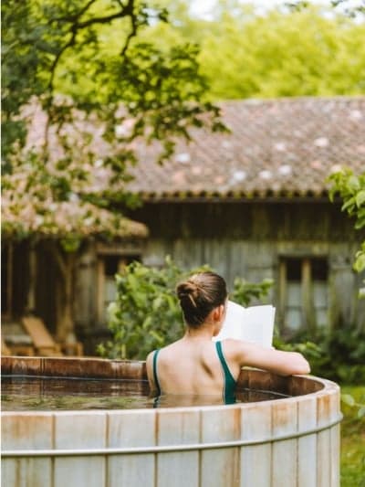 A guest at the cure, alone with a book in an outdoor wooden bath, the property's roofline beyond.
