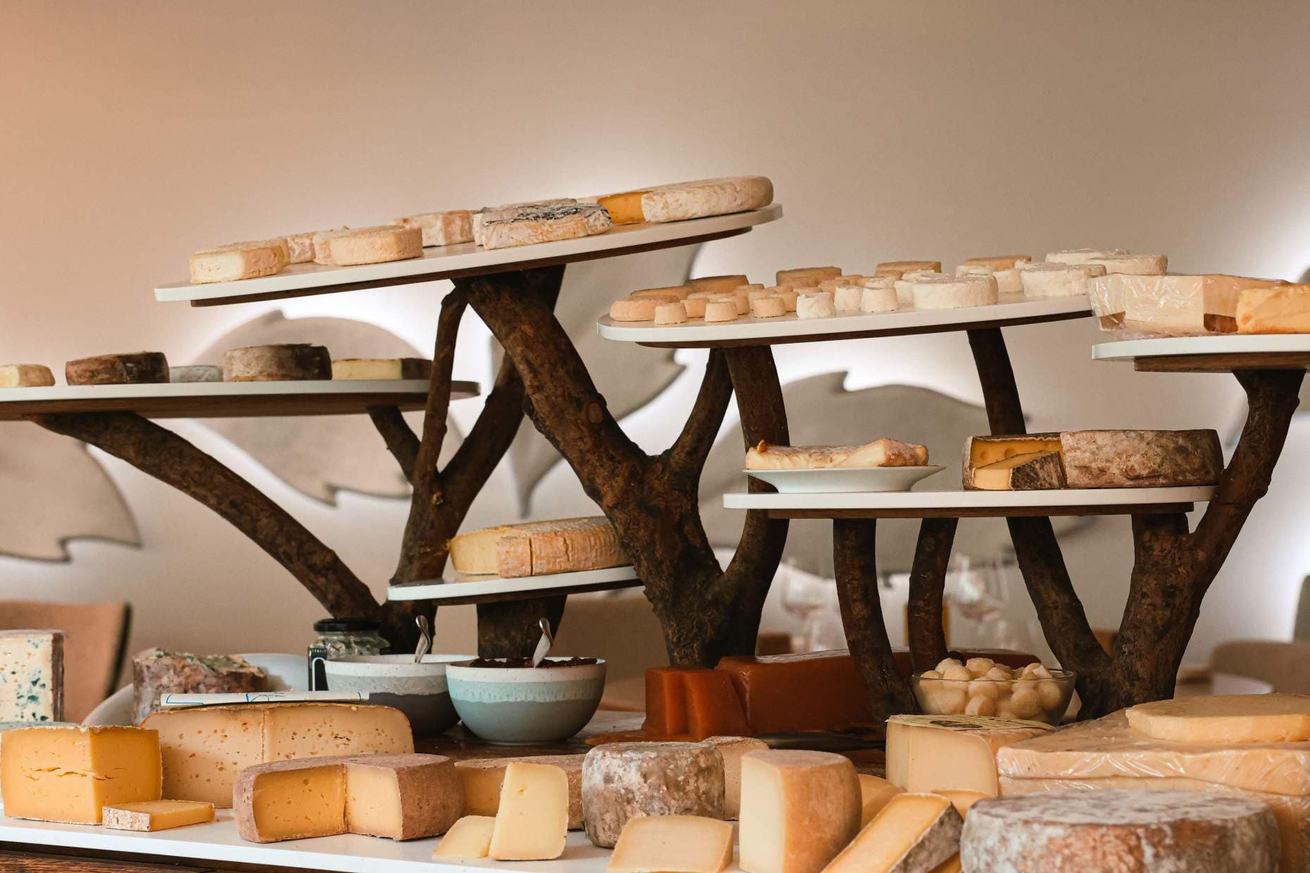 The cheese trolley at Auberge du Vieux Puits — three tiers of pale rounds and ash-rinded wedges balanced on twisted-vine stands, a row of jam pots in front and the dining room beyond.