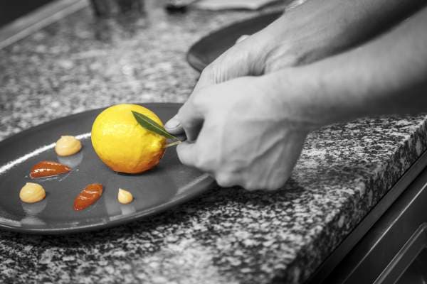 The chef's hand finishing a citron de Menton dessert in the pass — a yellow lemon trompe-l'œil on a grey plate, three caramel and ivory droplets in a half-circle, the photograph desaturated except for the lemon.