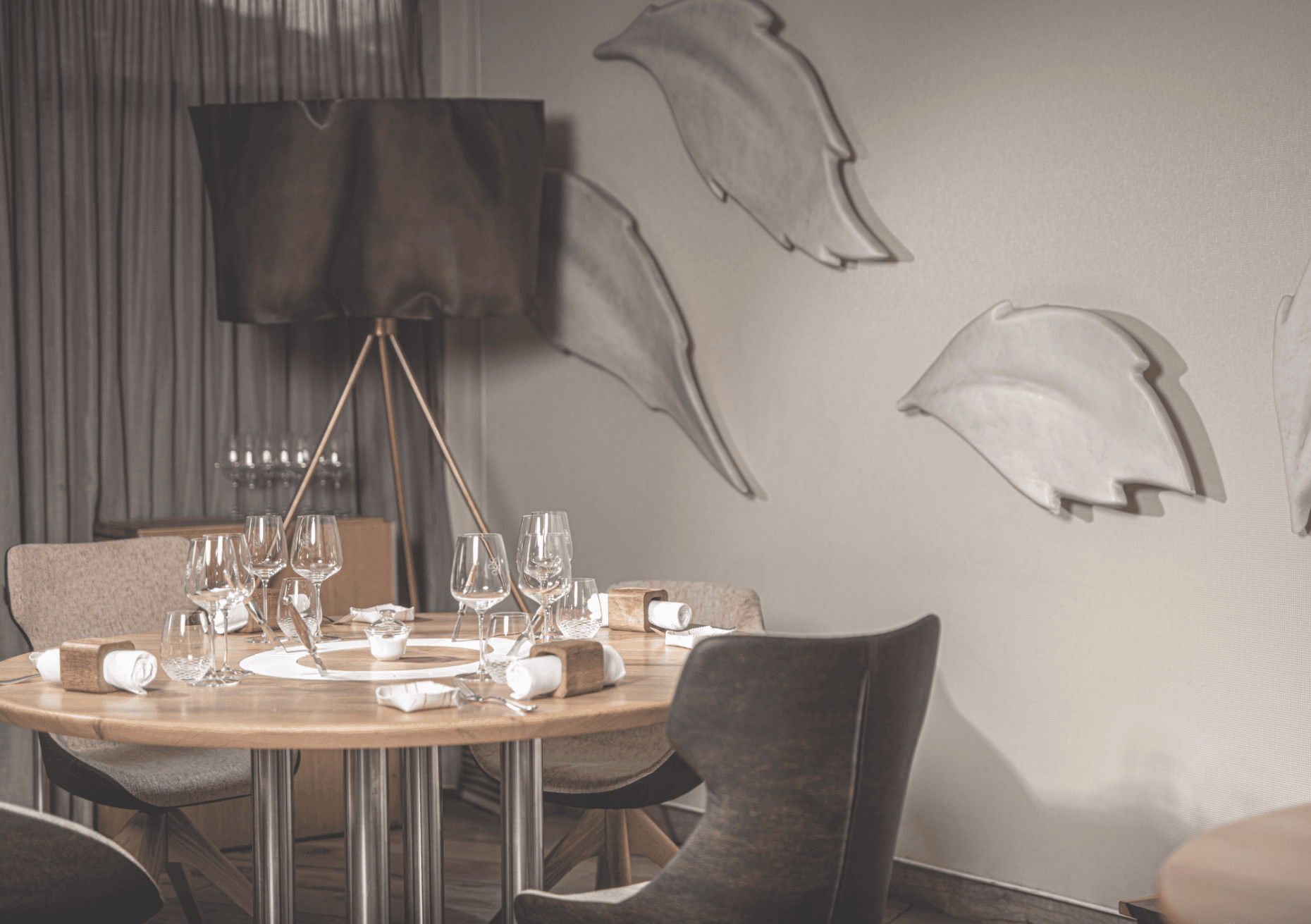 A round oak table set for two against the dining-room wall — two glasses, a wooden napkin ring, an amuse-bouche under a pale linen-leaf sculpture lit from behind.