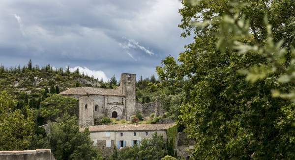 A view of Fontjoncouse from the road below — a Romanesque chapel with a square bell tower, a row of stone houses with terracotta roofs, the Corbières hills rising under a high cloud.