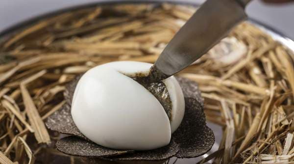 A close still life — a poached egg in its shell on a bed of dry straw, a thin paring knife setting into the shoulder of the egg to a smear of black truffle.