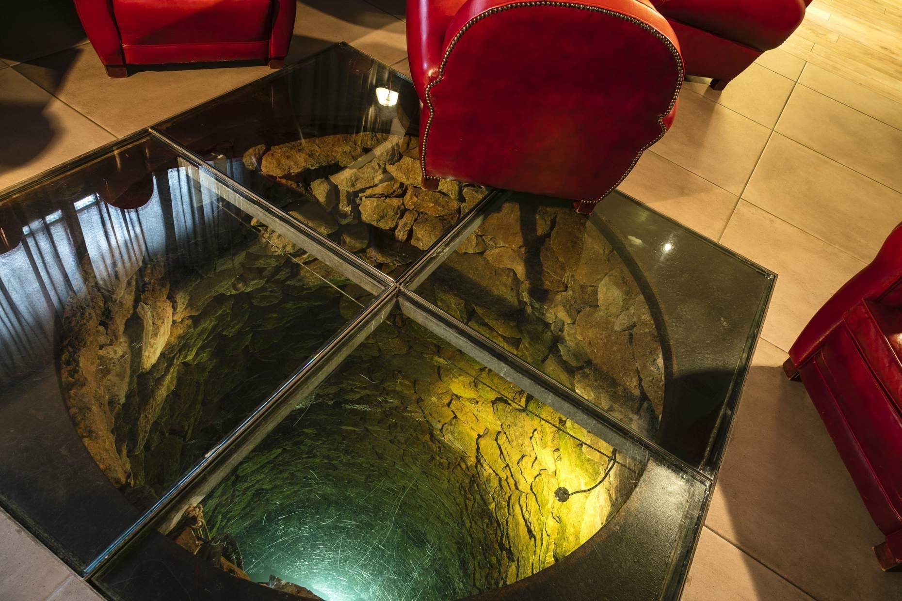 The old village well seen from above through a glass floor in the salon — green-lit stone shaft dropping into shadow, deep red leather armchairs gathered around the rim, terracotta tiles beyond.