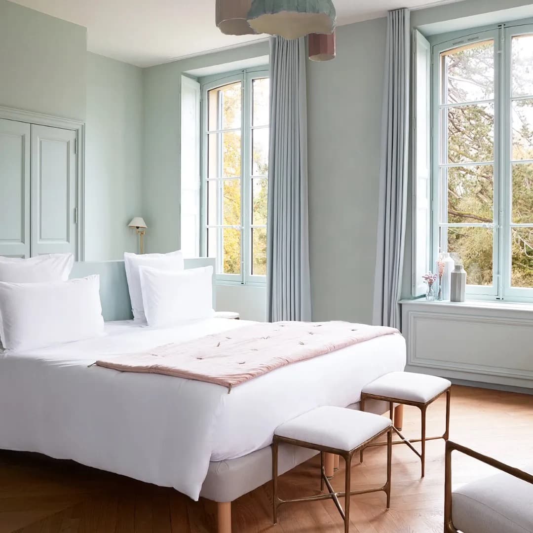 A pale-mint guest room with tall windows, white linen and a pink throw on the bed, the park visible beyond the panes.