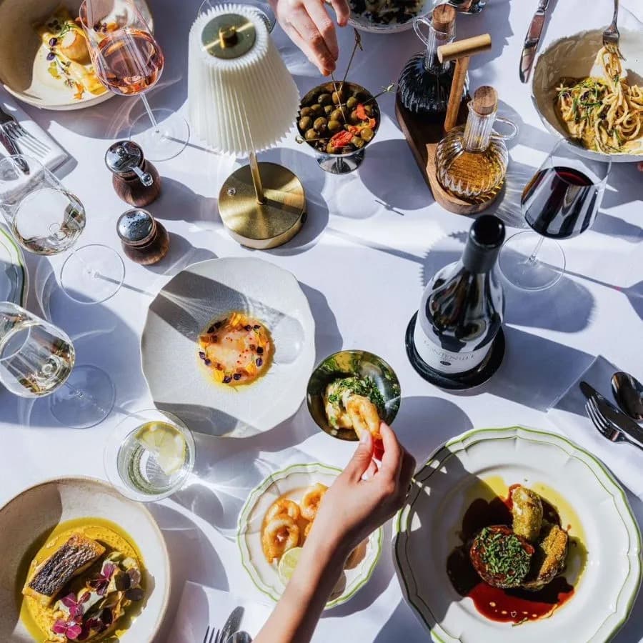A long white-linen table seen from above, plates of pasta and small dishes in midday sun, hands reaching across the cloth.