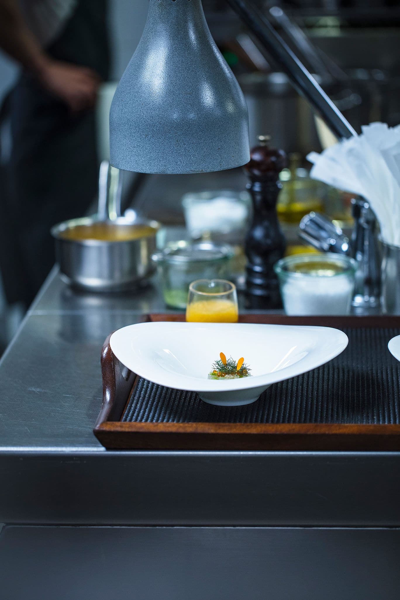 A single white plate set on a wood tray under a steel heat lamp at the pass — a small composition of dill and citrus on a smear of dark sauce, a glass jug and a pepper mill behind.