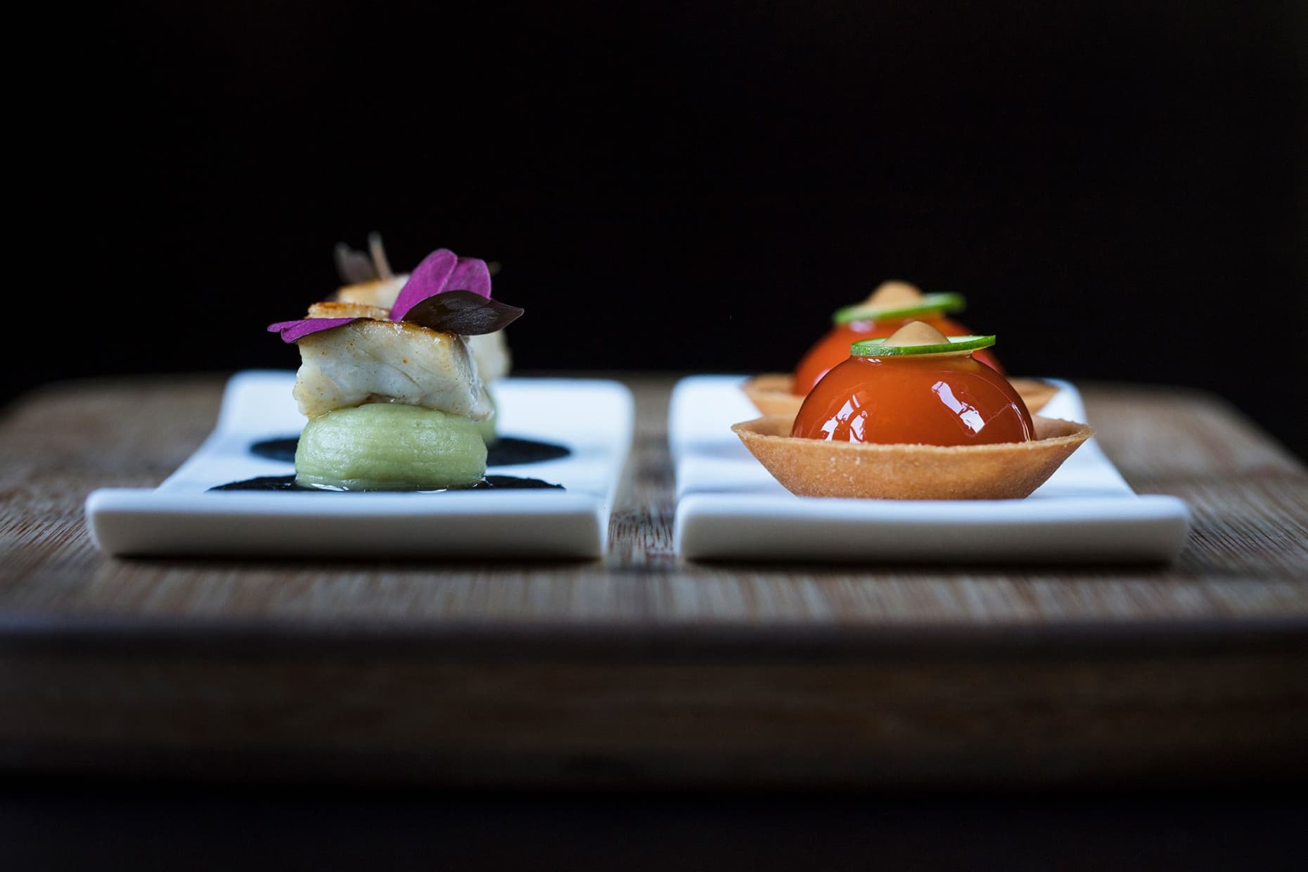 Two amuse-bouche on small white plates against a black background — a seared scallop on a green pea purée and a glossy red tomato confit in a thin pastry shell.
