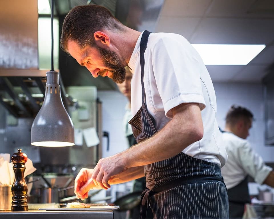 Michaël Arnoult in profile under a heat lamp at the pass — focused, plating a single dish, his black-striped apron and white jacket lit from the side.