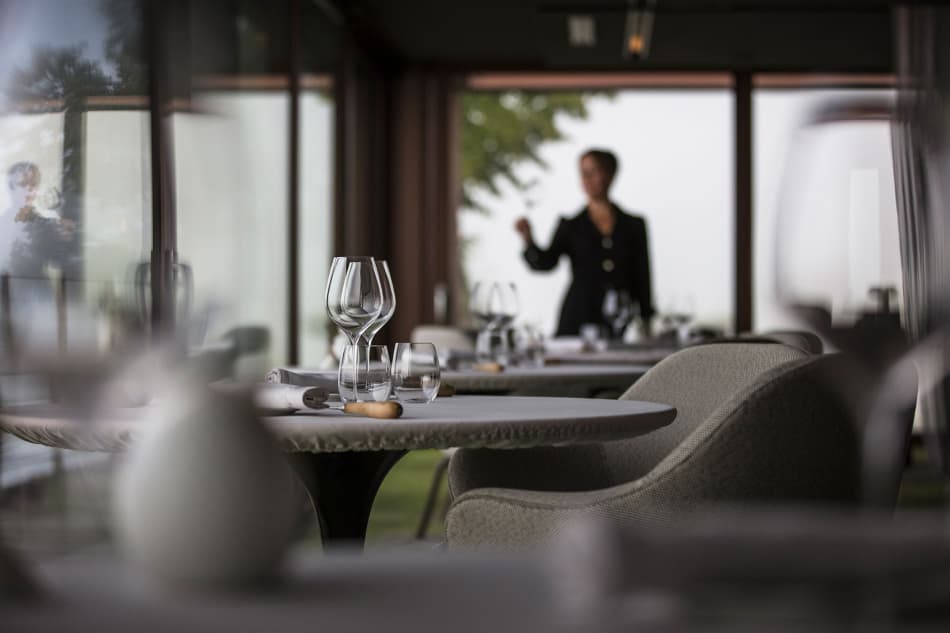 The dining room interior — a sommelier in a dark jacket silhouetted between rows of polished wine glasses, a low table with crystal and a small bread roll, soft afternoon light through full-height windows.