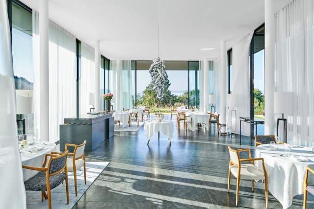 The Louison dining room at Villa La Coste — white tables, a hanging sculpture, glass walls open onto the gardens.