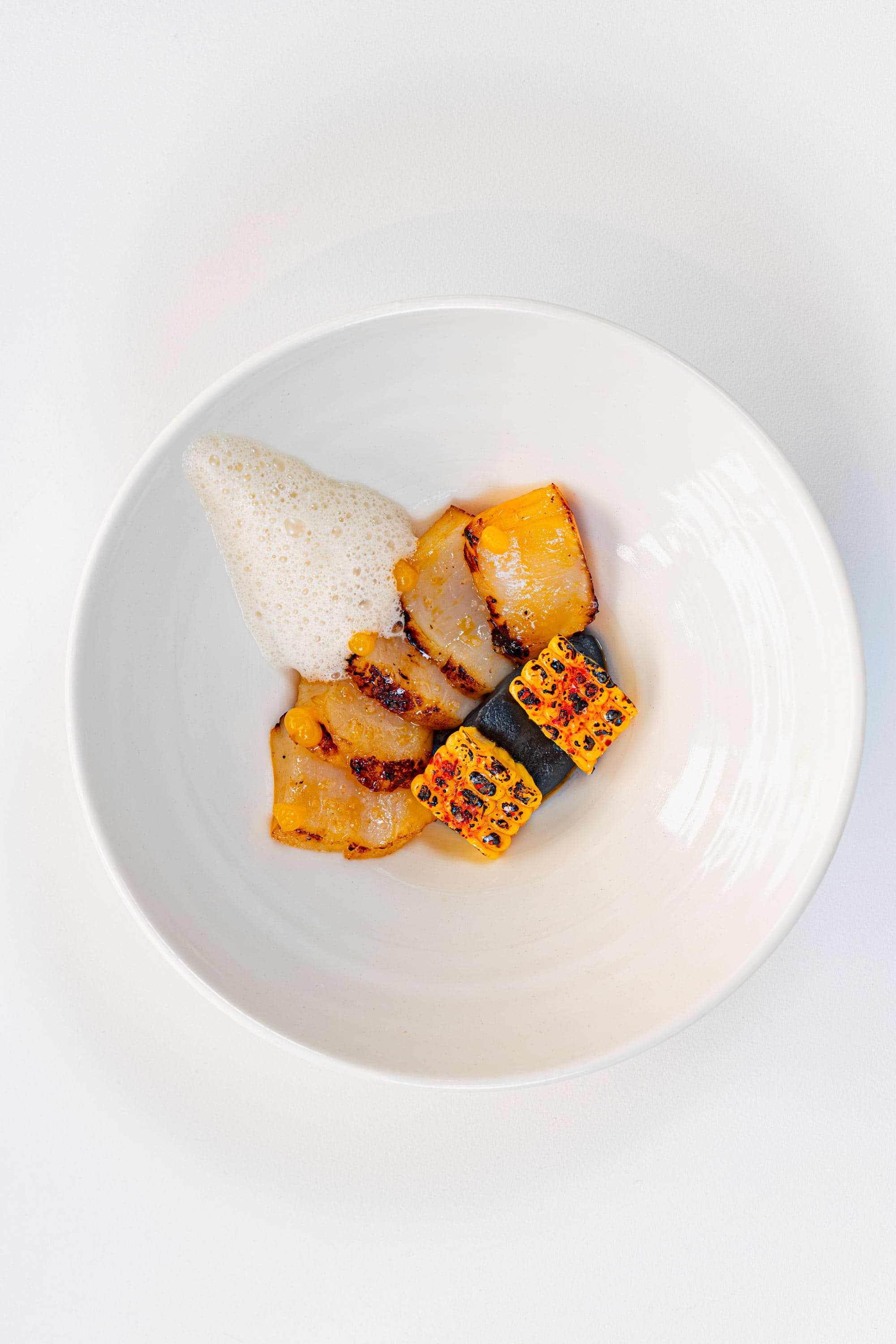 A single white bowl from above — slices of seared scallop, a cube of charred corn over a black quenelle and a soft white foam, a single scallop placed apart on the rim.