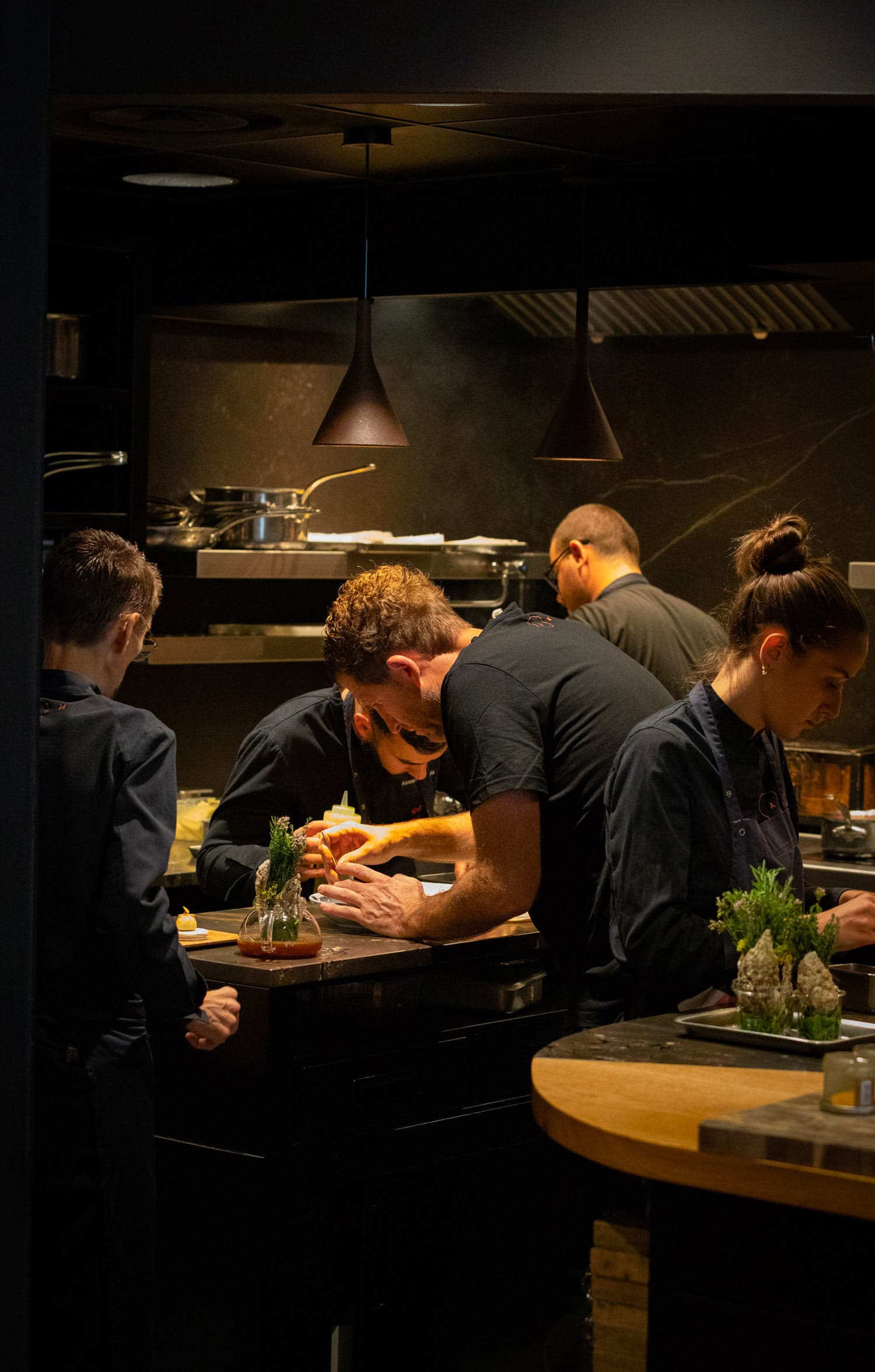 The brigade at the pass in profile — three cooks in dark jackets bent over their boards, low pendant lamps, rosemary and thyme on a wooden block in the foreground.