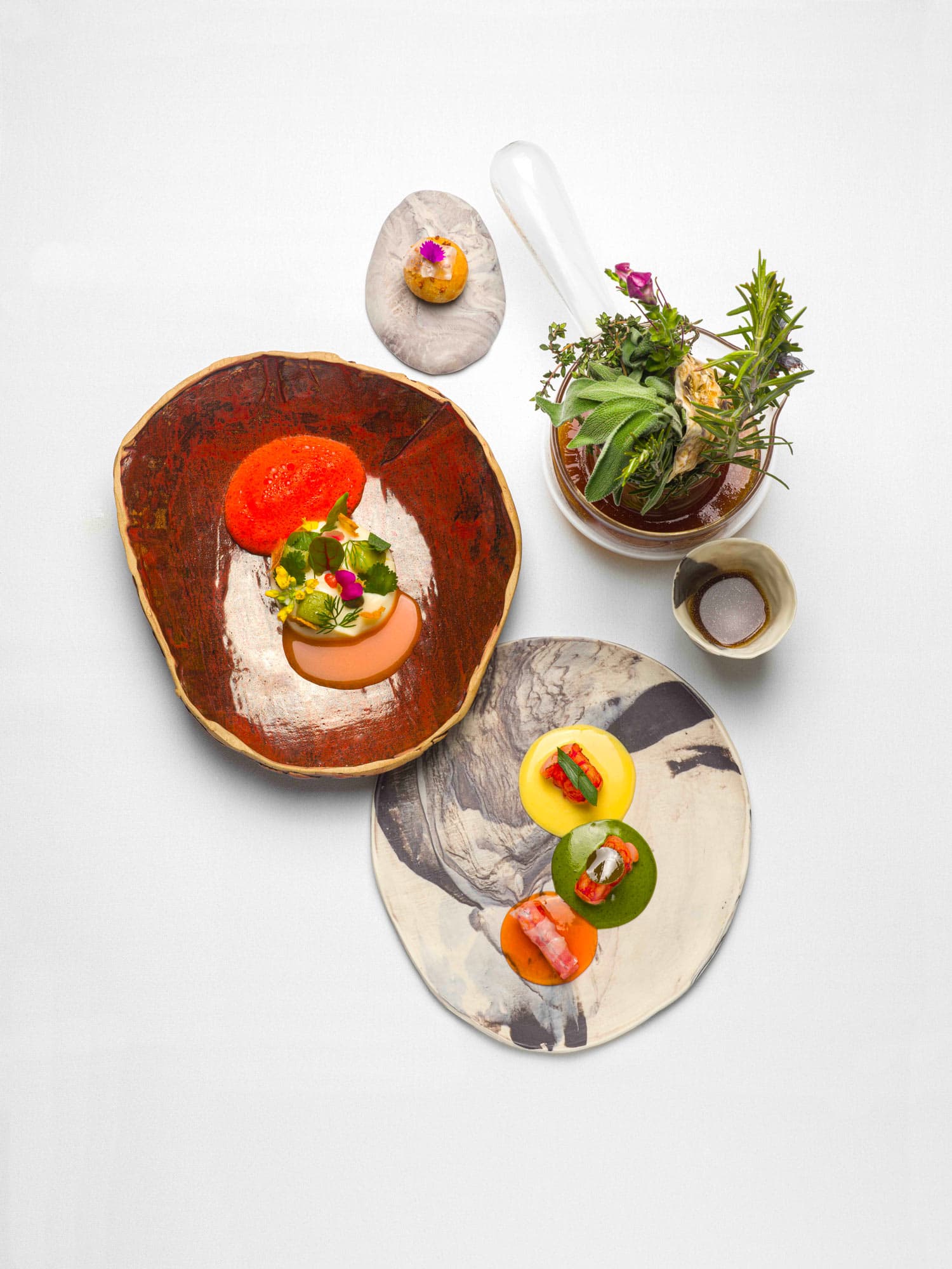 An overhead arrangement — a deep terracotta plate with two small bites at its centre, a glass mortar of fresh herbs with a long pestle, a marble dish of three round bouchées and a small ceramic of salt.