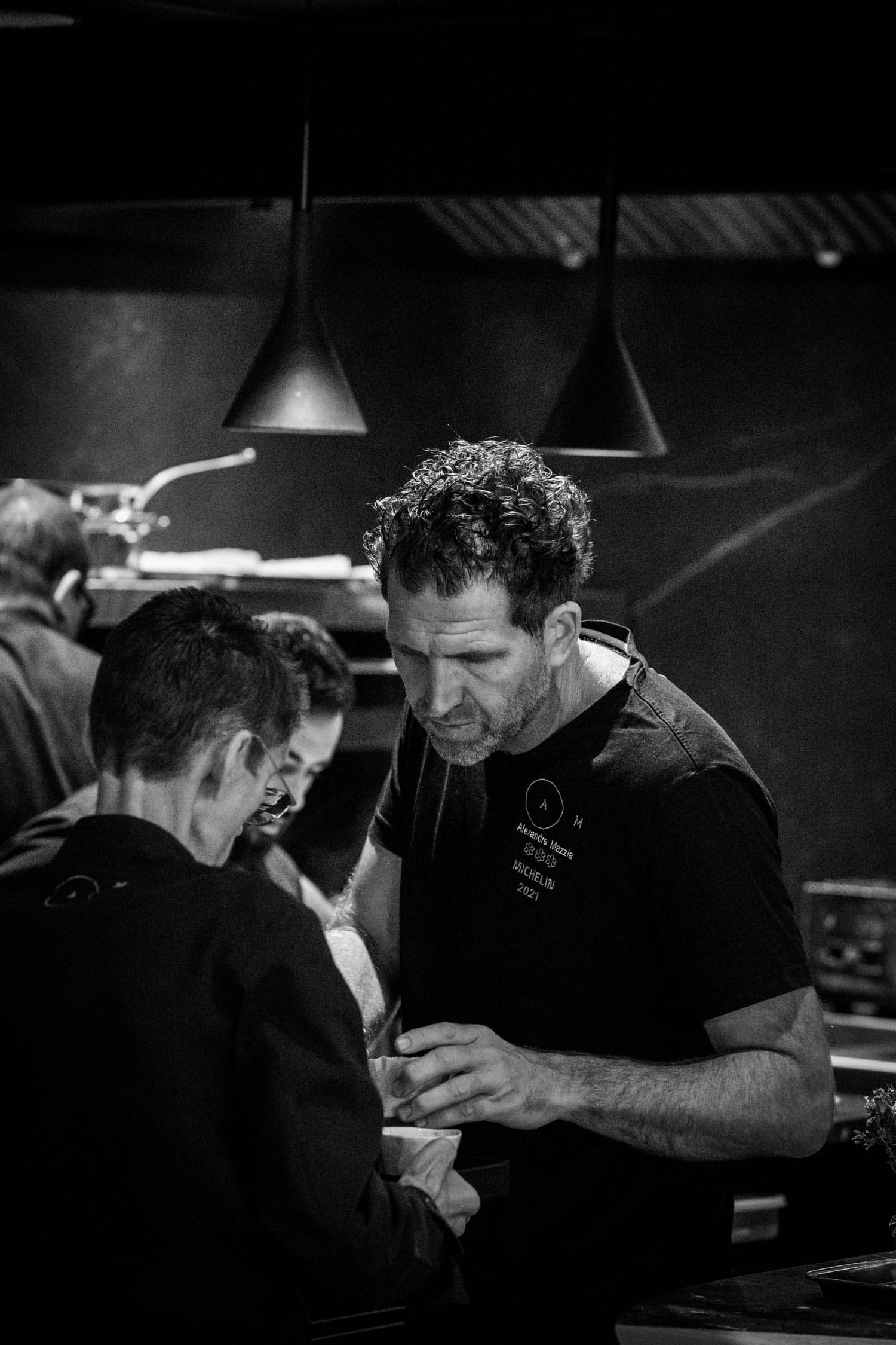 A black-and-white portrait of Alexandre Mazzia at the pass — leaning over a finished plate, curls in profile, a black t-shirt with the letters A and M and three Michelin stars at the chest, two pendant lamps overhead.