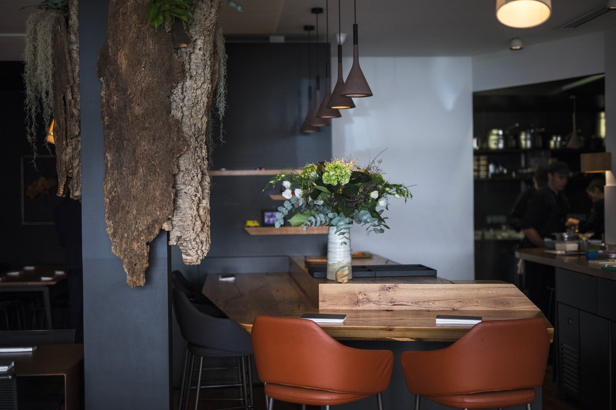 A close-in view of the dining room — a panel of weathered cork bark trailing strands of moss, a row of bronze pendant lights, two leather armchairs at a wooden counter, an arrangement of greenery on the table.