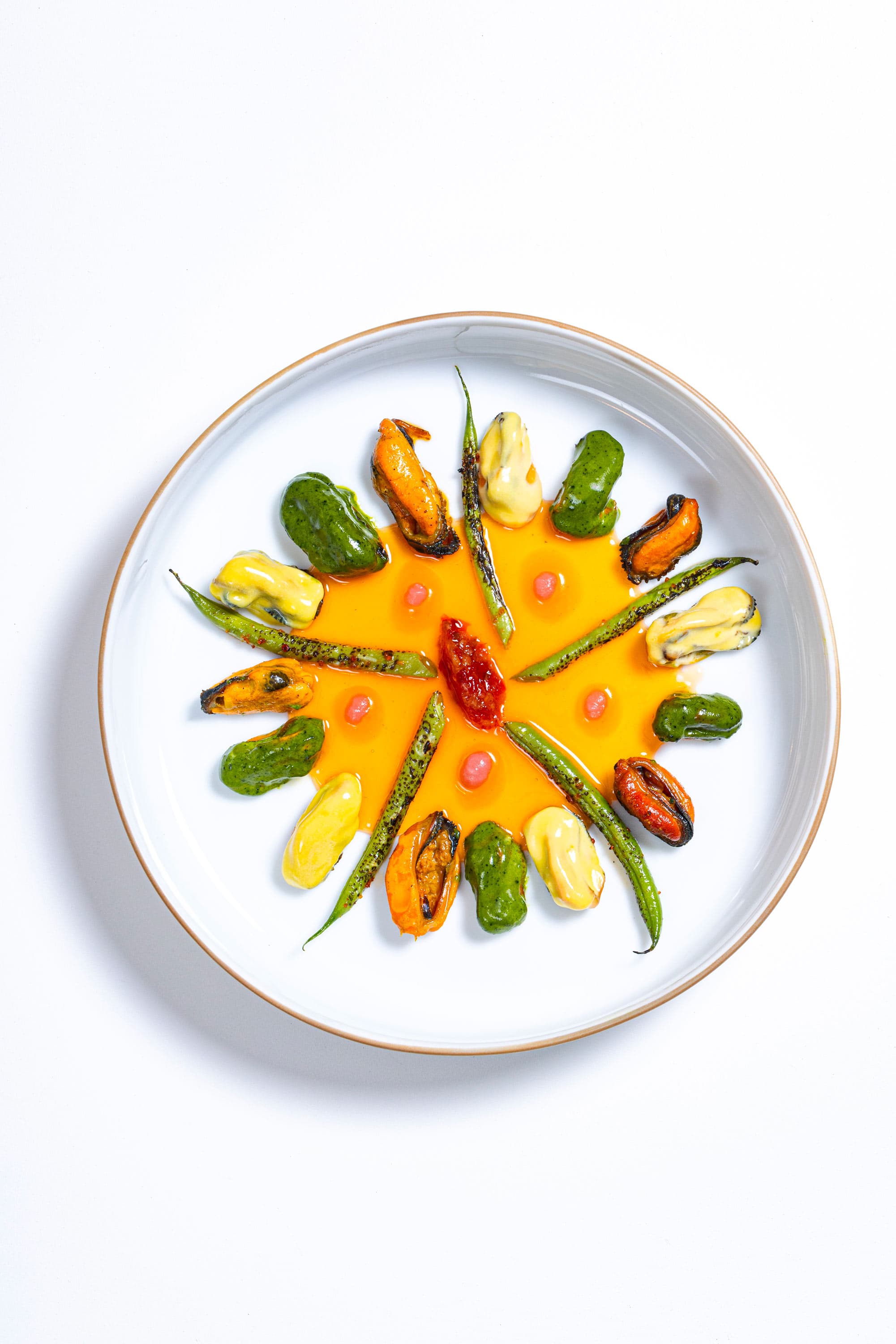 A signature plate from above — small mussels and pieces of grilled mackerel arranged radially with green herbs and yellow sauce dots in a wide pool of saffron-orange emulsion on a white plate.