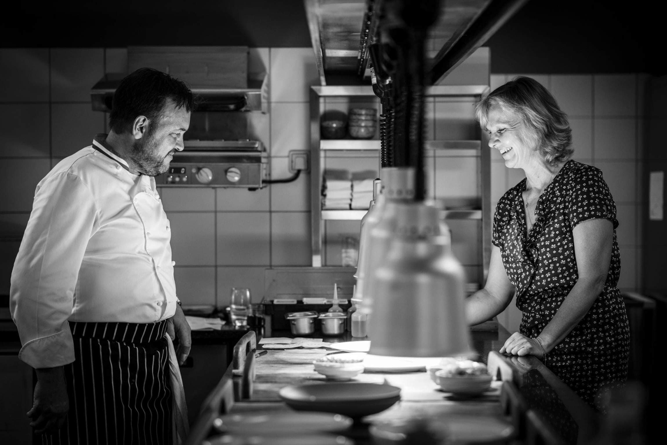 Black-and-white pass shot — Emmanuel Renaut in white jacket and pinstriped apron, standing at the kitchen line under a heat lamp, Kristine Renaut at the far end laughing in a print dress, plates and small bowls between them.