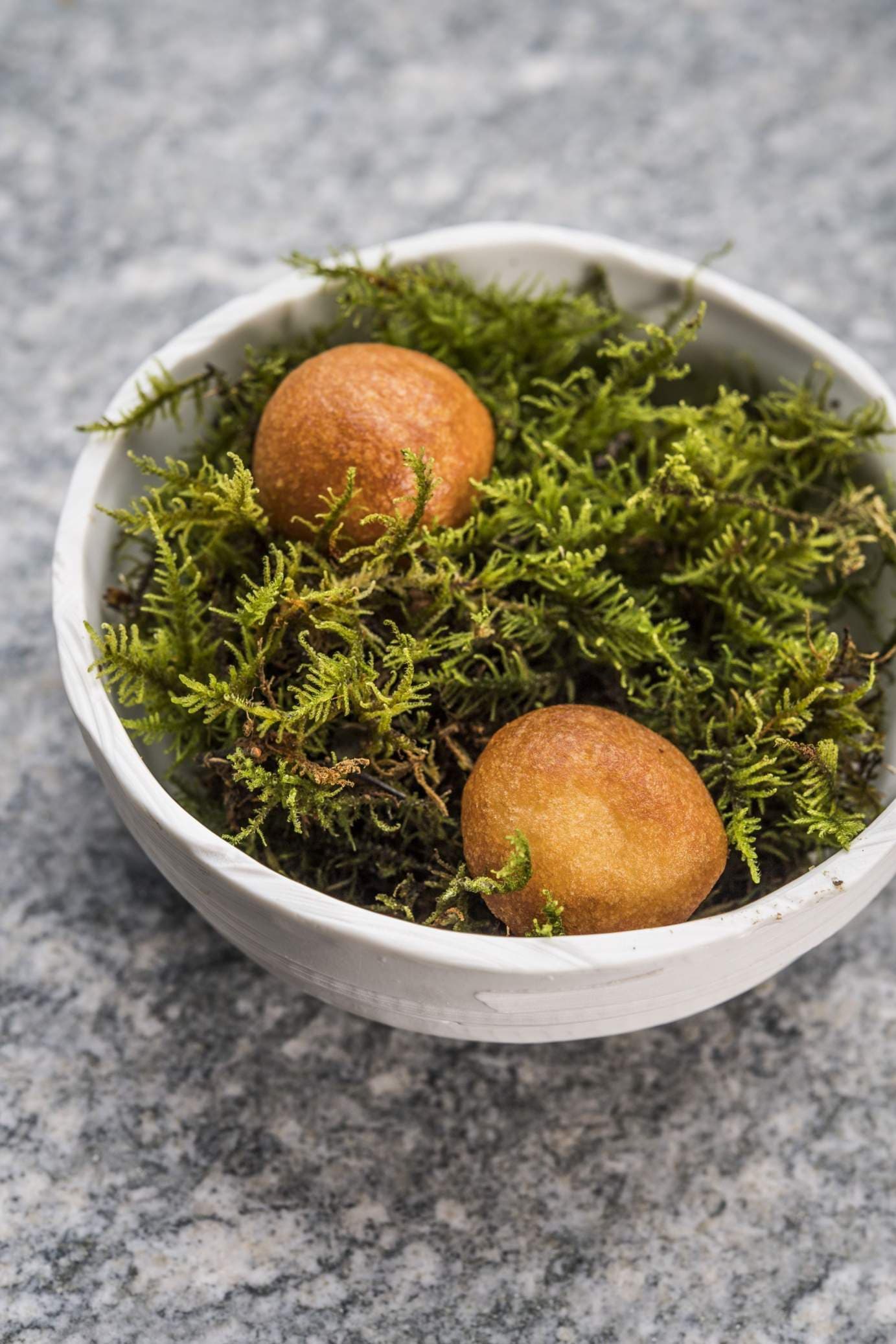 Two golden-brown fritters set in a nest of bright green moss in a small white bowl on grey stone — a foraged-feeling amuse-bouche.