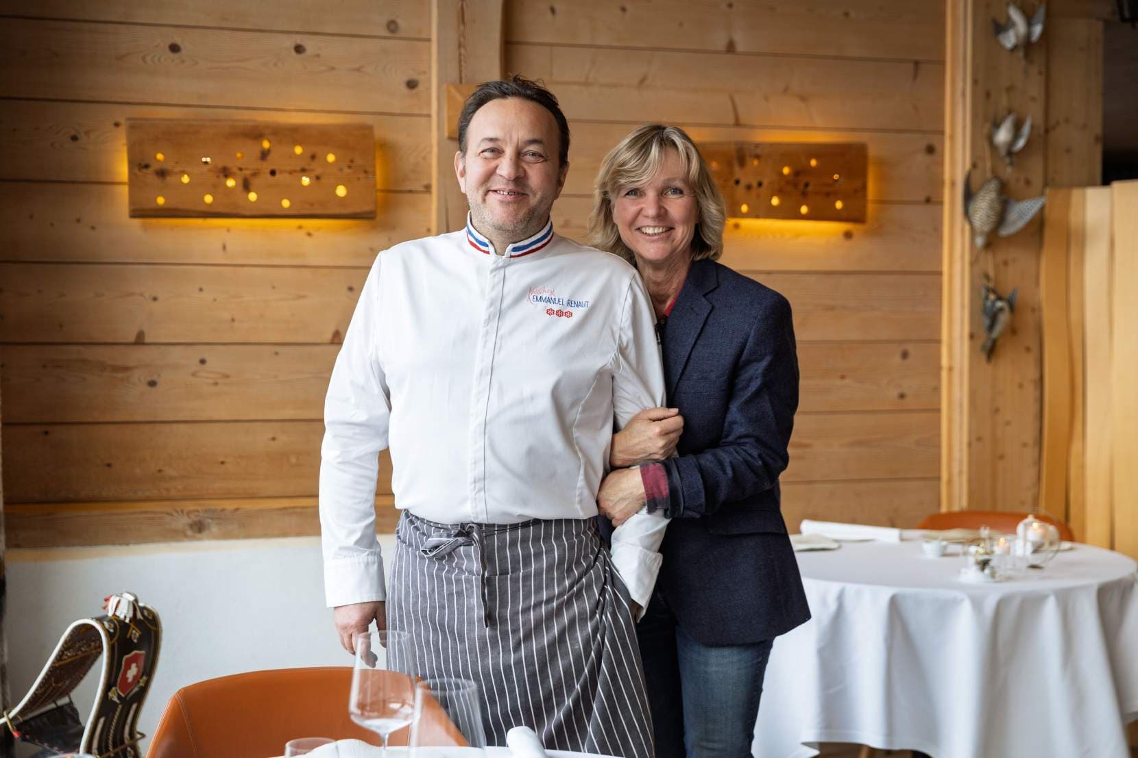 Emmanuel and Kristine Renaut in the dining room — the chef in a white jacket with the red-white-blue MOF collar embroidered with his name, his wife in a navy blazer at his shoulder, both looking off-camera, two iron birds on the wood-clad wall behind them.