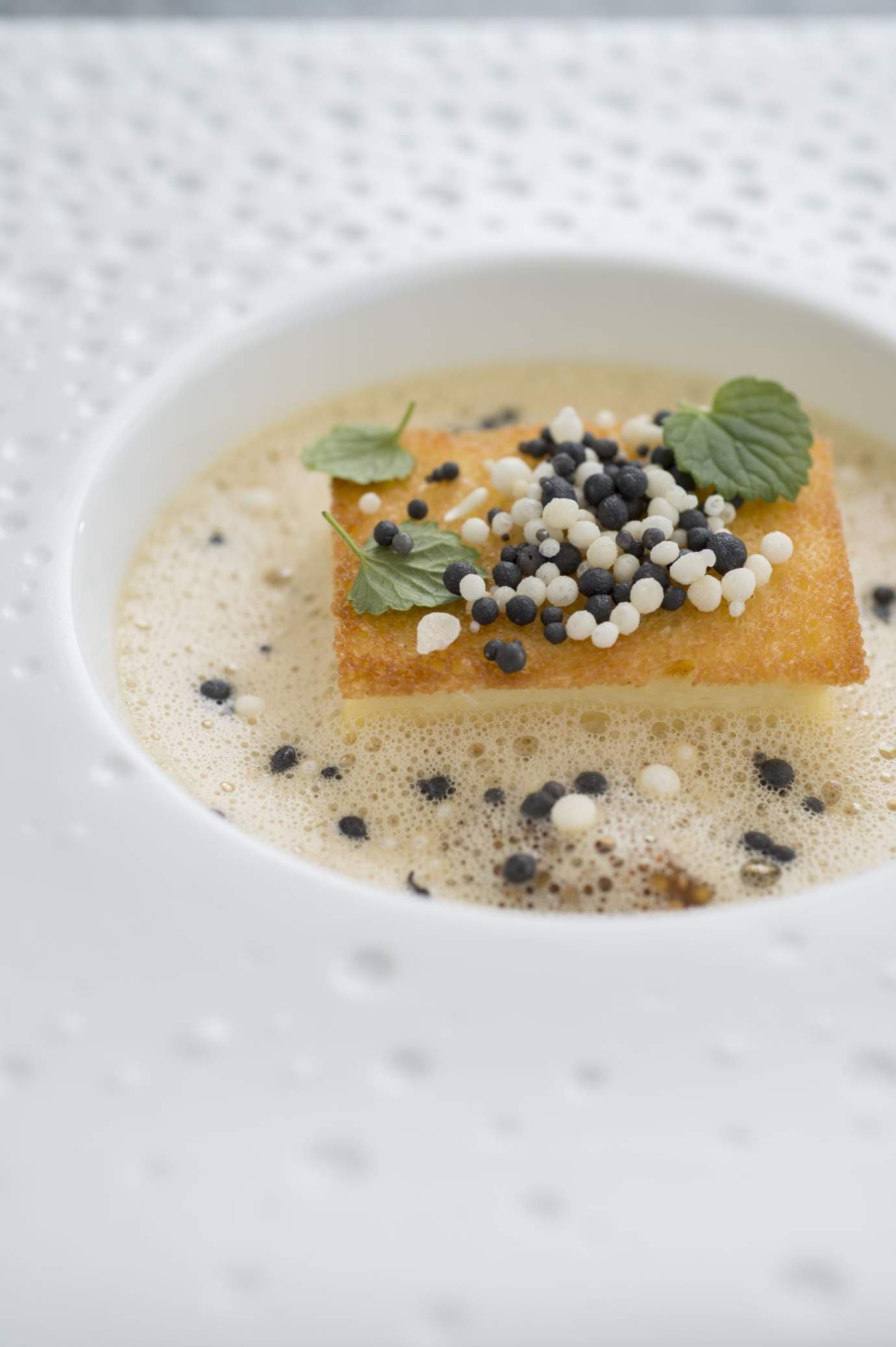 A close overhead of a white porcelain bowl with a pale square of pike biscuit topped with black and white caviar pearls and two small green leaves, set in a foaming watercress velouté.
