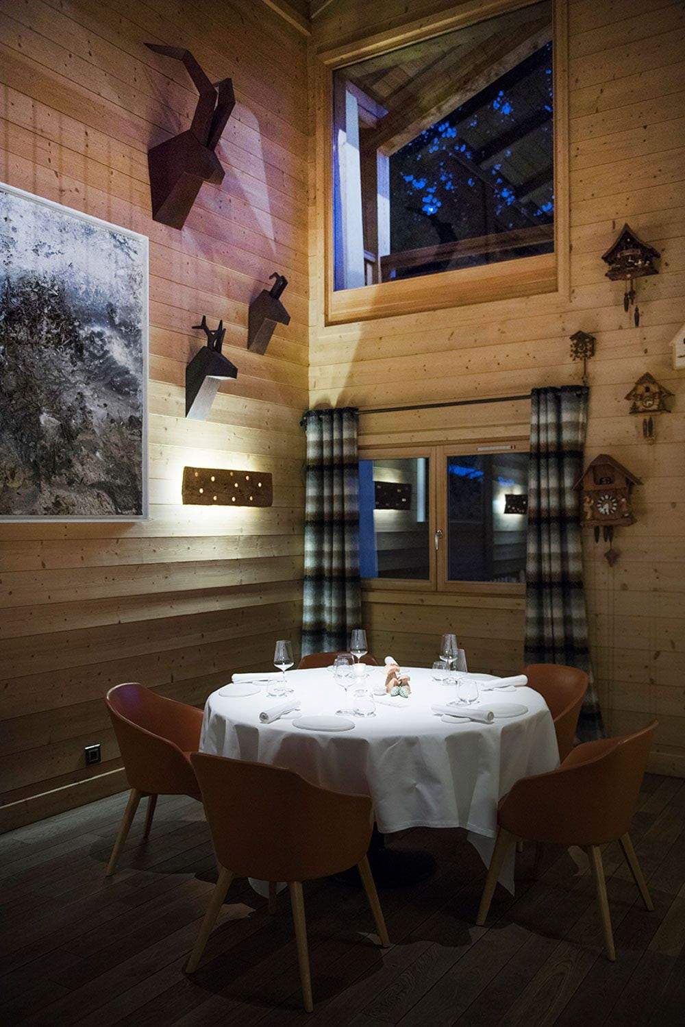 A small chalet dining alcove — a single round table for four under a tall window, pine plank walls hung with iron chamois sculptures, a black-and-white mountain photograph and three cuckoo clocks, the table laid in white linen with stemware.