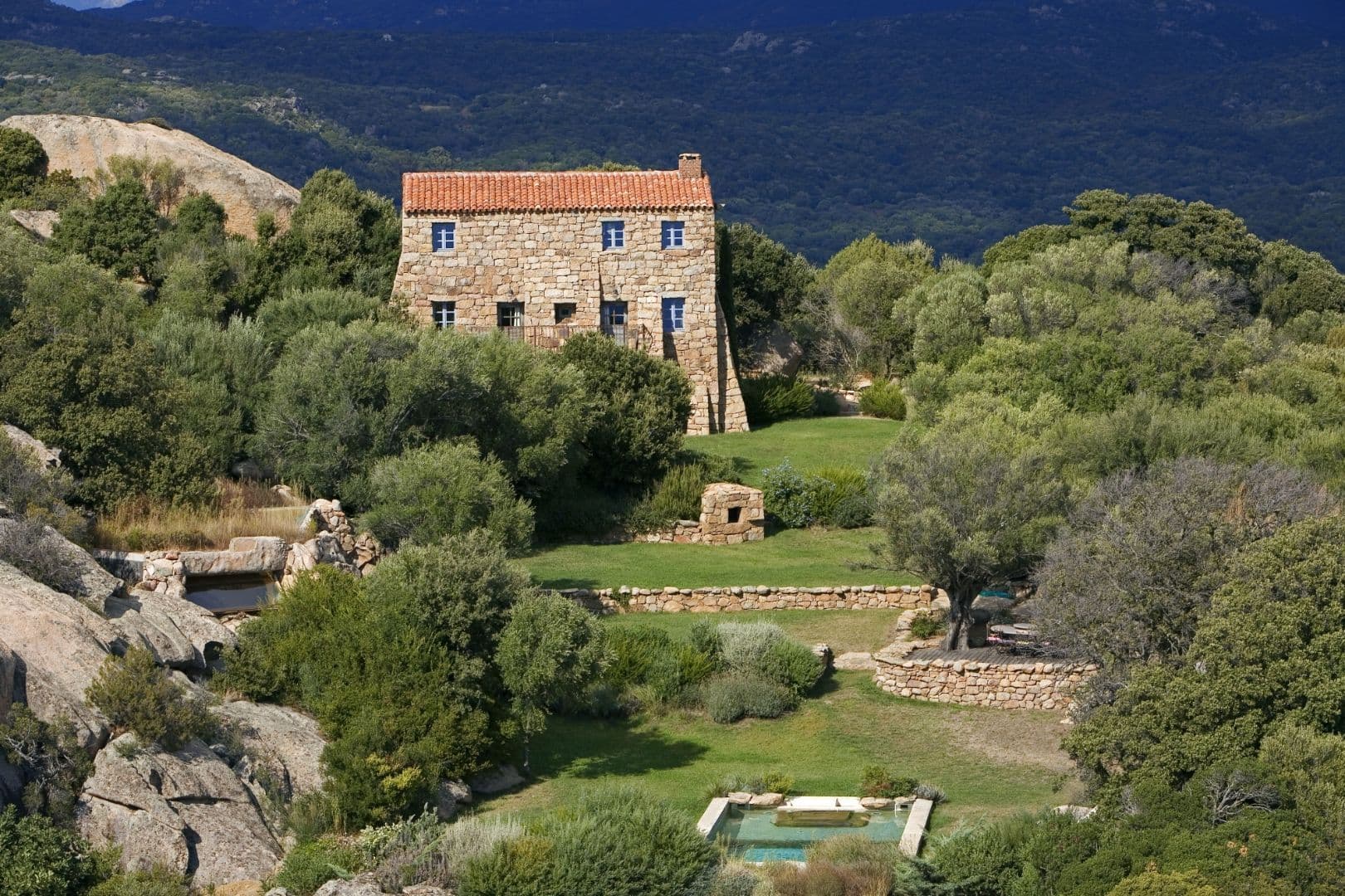 A restored stone bergerie set against a granite outcrop, mountains drifting away behind, a small swimming pool tucked into the garden below.