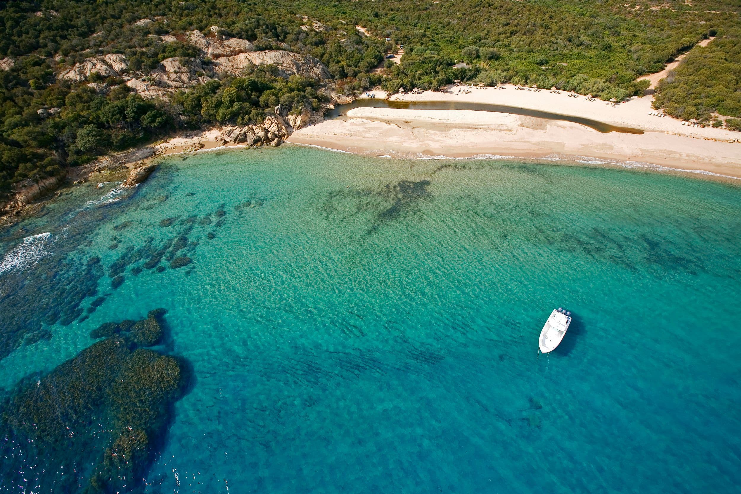 An aerial view of one of the estate's coves — turquoise water over granite shelves, a single white boat at anchor, the maquis closing in to the sand.