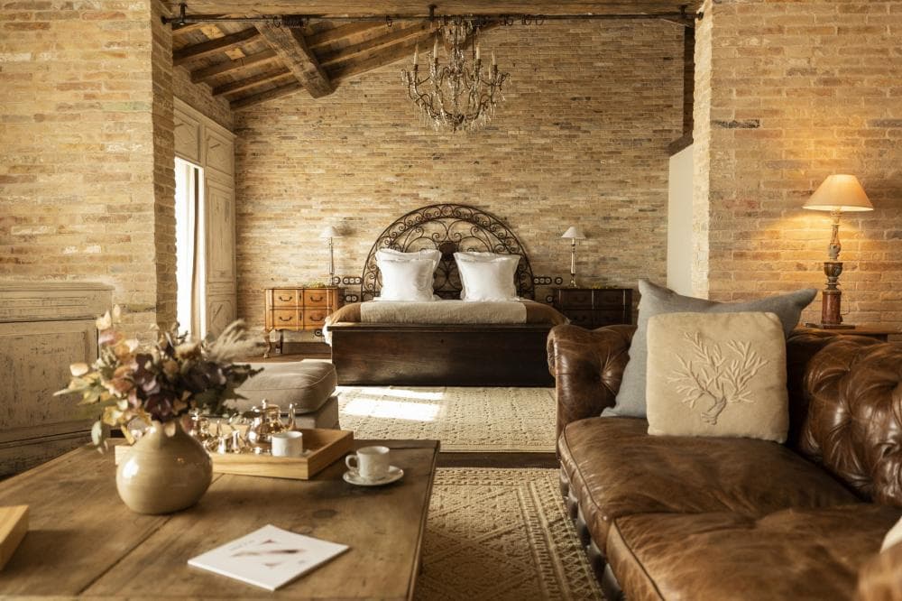 The interior of a restored bergerie — pale brick walls, exposed beams, a forged-iron headboard, a leather chesterfield turned to a dressed bed under a small chandelier.