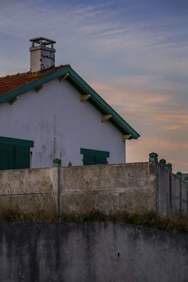 The Vendée gable end of an island cottage at sunset — white walls, terracotta tile, sea-green shutters and a cinder-block sea wall in front.