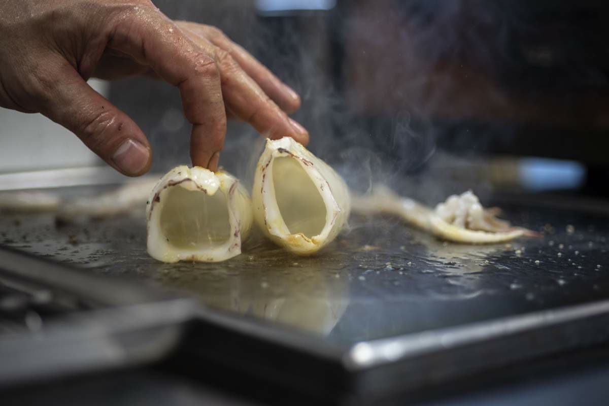 A close hand on the plancha, lifting a translucent shell of seasoned squid as steam rises across the steel.