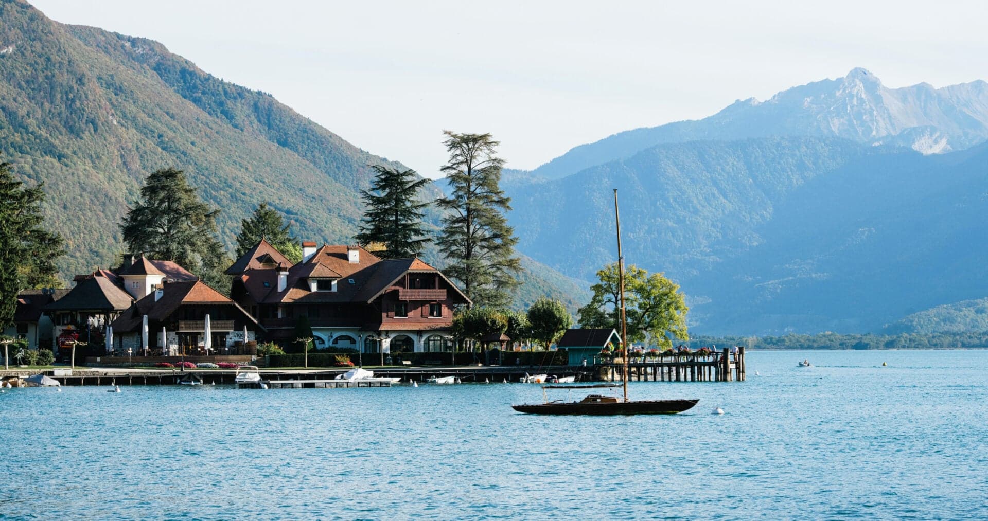 The auberge from the lake at midday, a small sailboat moored in front of the chalet roofs.