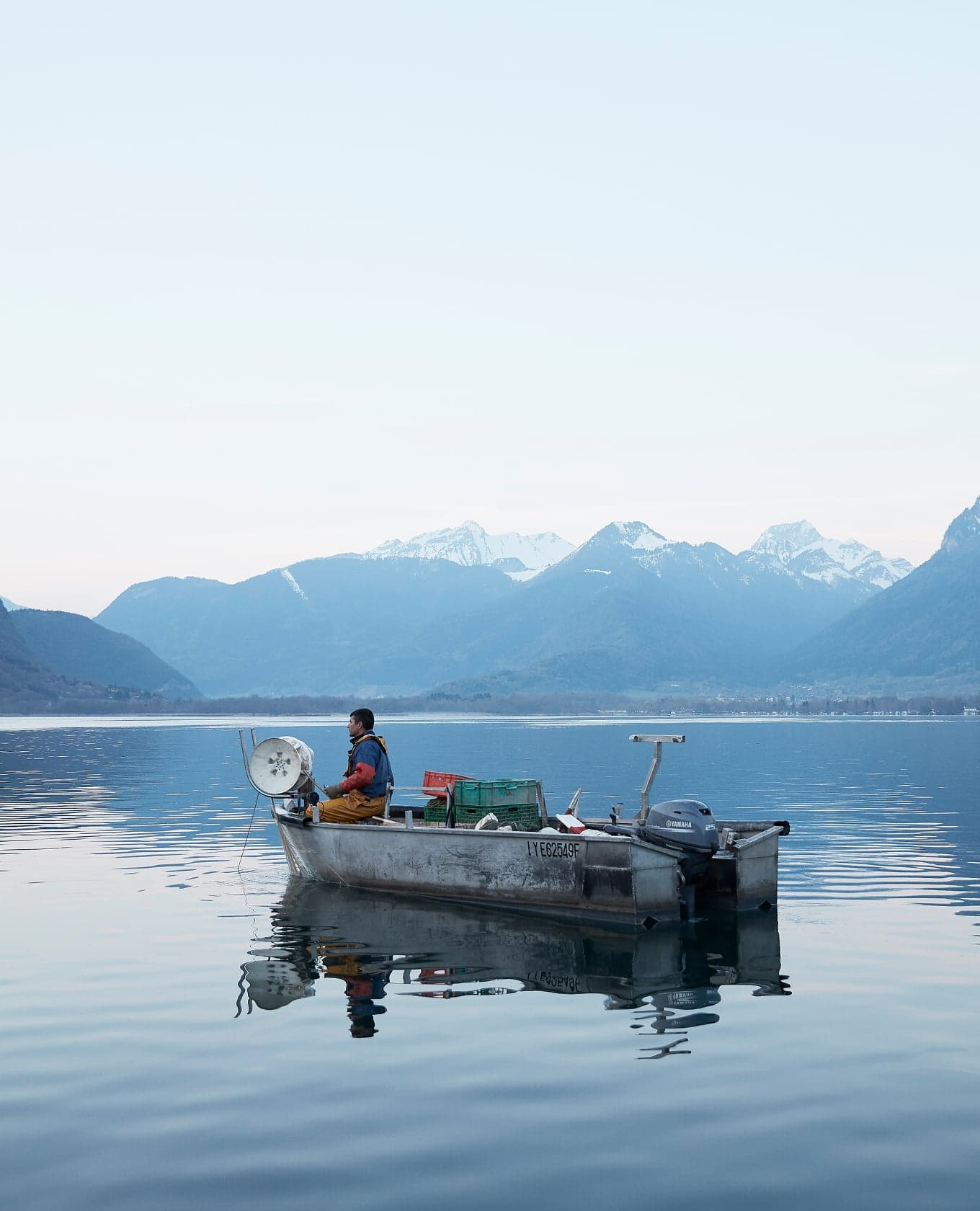 A lone fisherman on the lake at first light, the snow-capped alps reflected in still water.