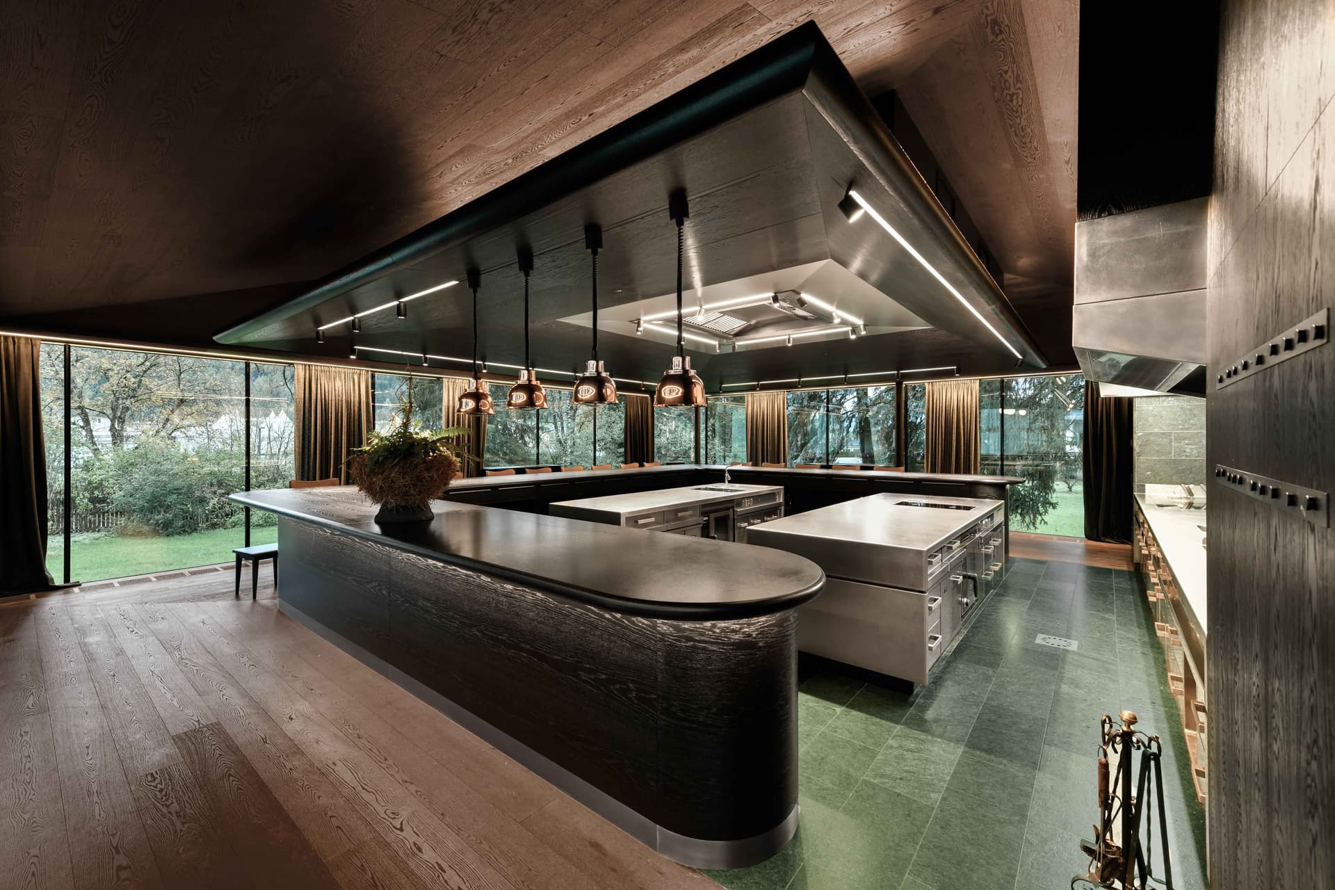 The open-kitchen pavilion at Atelier Moessmer — a curved black-oak counter wrapping a steel pass, copper-bell pendant lamps in a row, full-height glazing onto the green Pusteria garden behind.