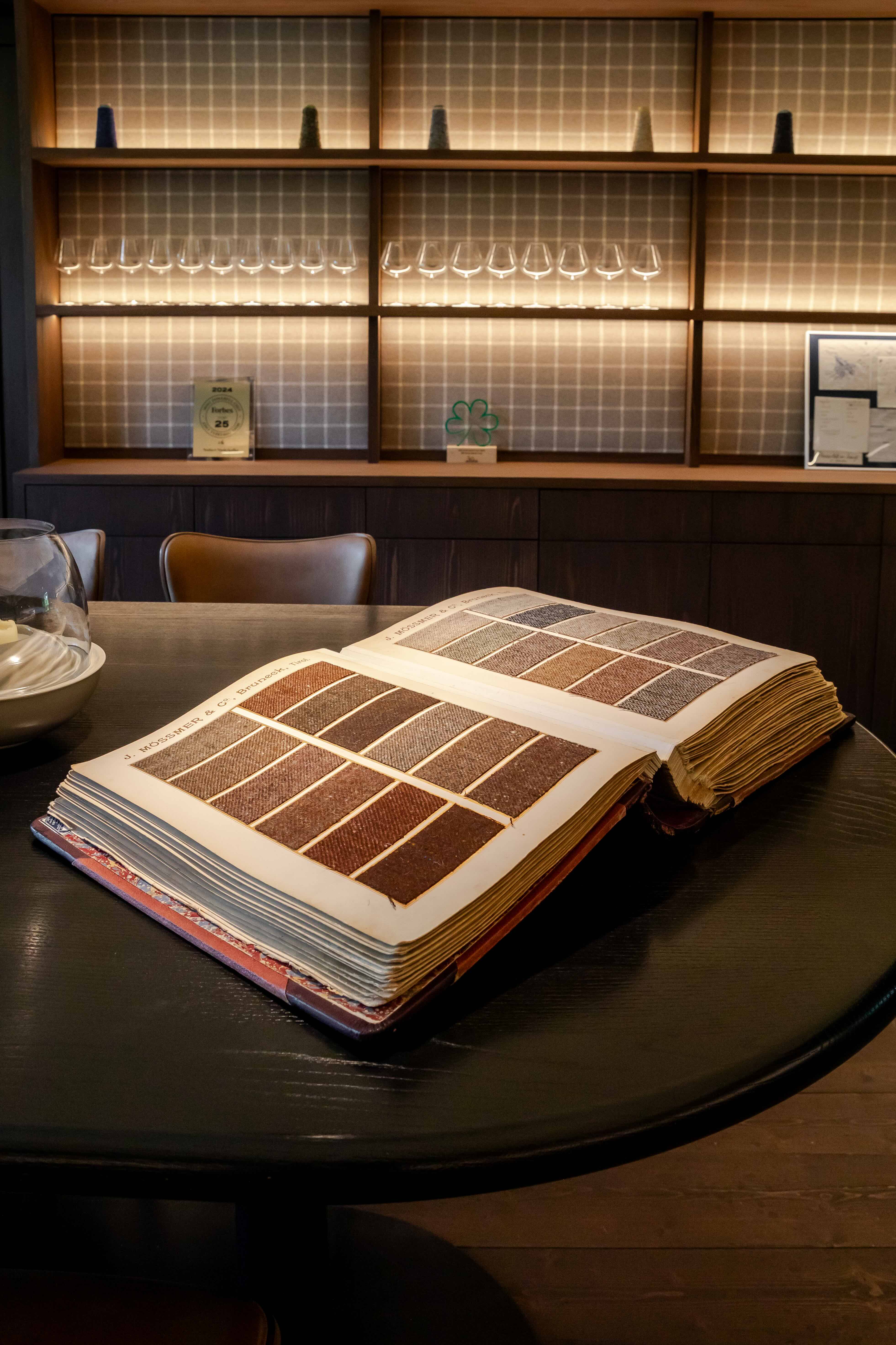 A Moessmer wool sample book opened on a black table — page on page of small woven swatches in greys, browns and reds, a backlit shelf of stemware behind in soft tile light.
