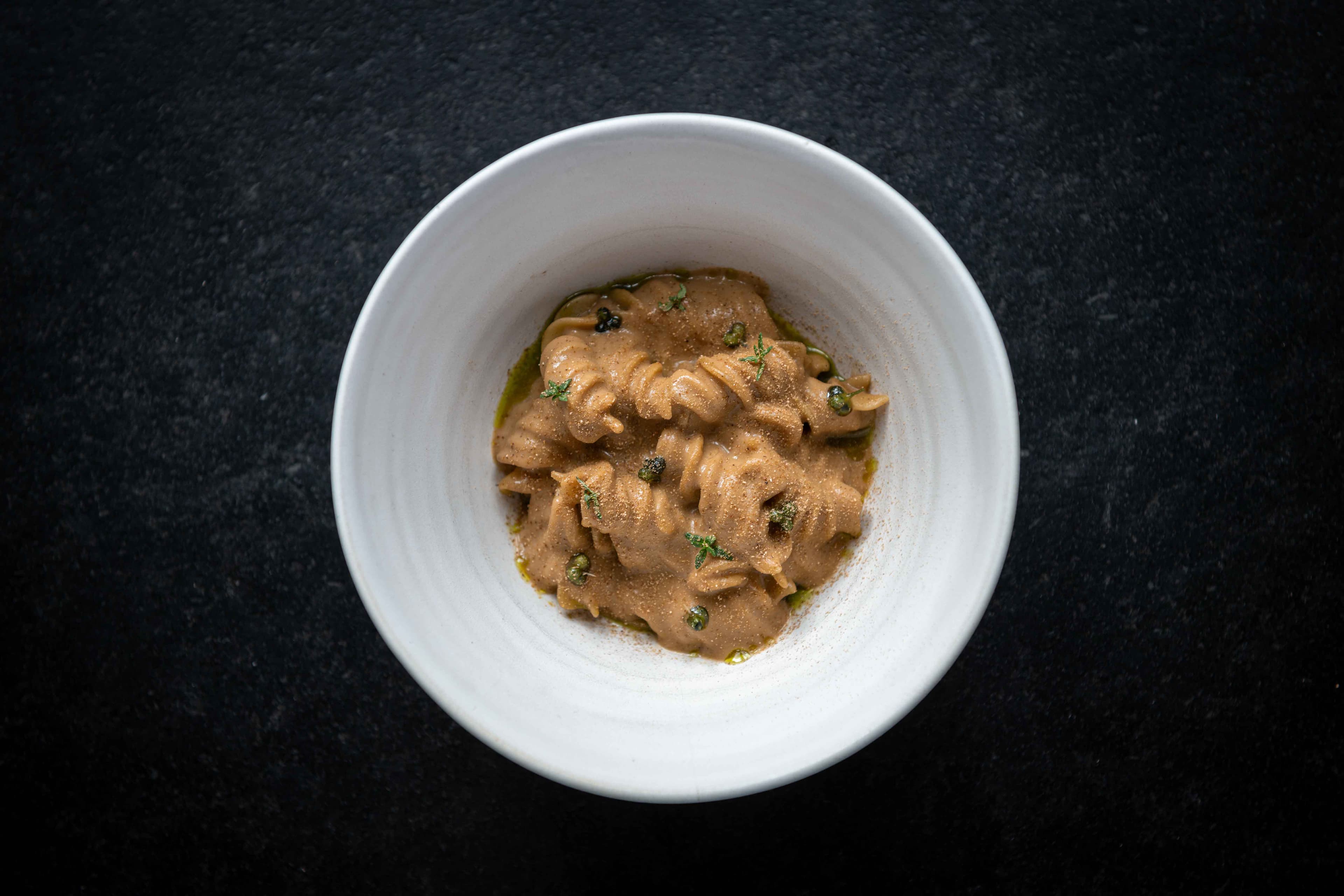 A pale ceramic bowl seen from above on a slate table — twists of fusillone pasta in a glossy fermented mushroom and koji cream, fine green herbs scattered on top.