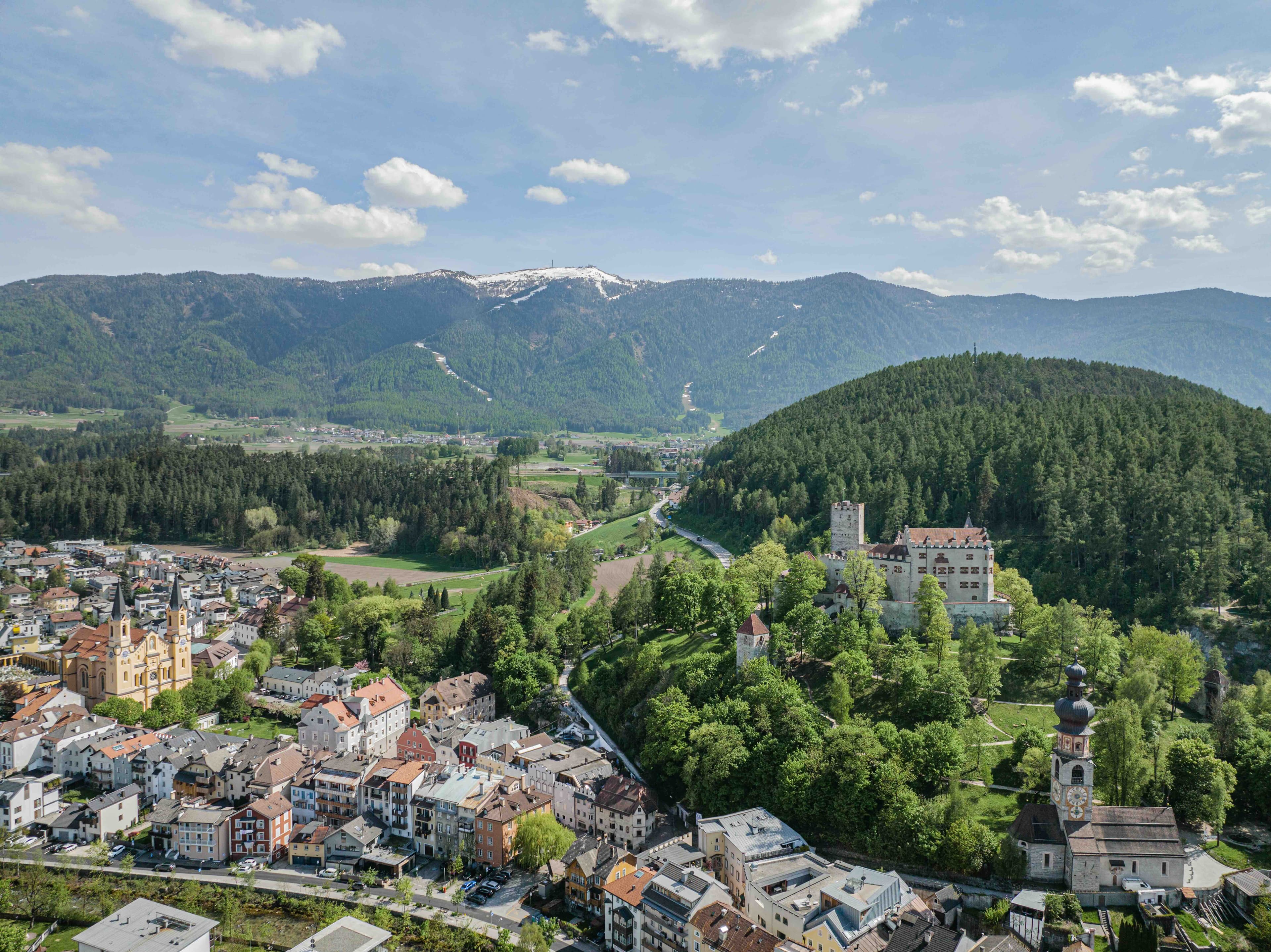 An aerial of Brunico in spring — terracotta rooftops of the old town along the Rienza river, Bruneck Castle on its wooded mound to the right, the snow-tipped ridge of Plan de Corones beyond.