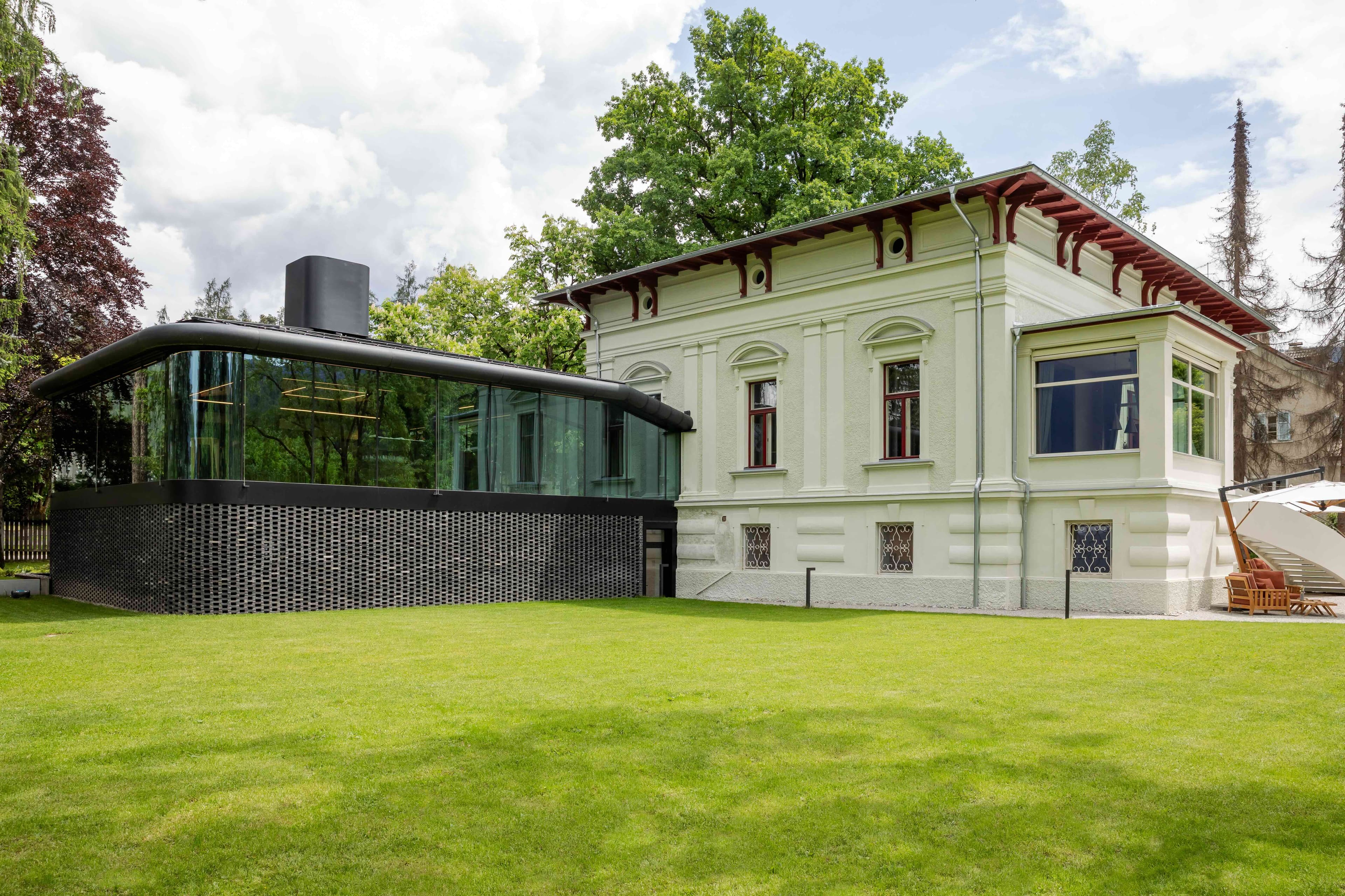 The Moessmer manor in summer — a pale late-classical façade with terracotta-trimmed cornices, a black-clad glass kitchen pavilion grafted to its left wing, lawns and tall trees of the park around.