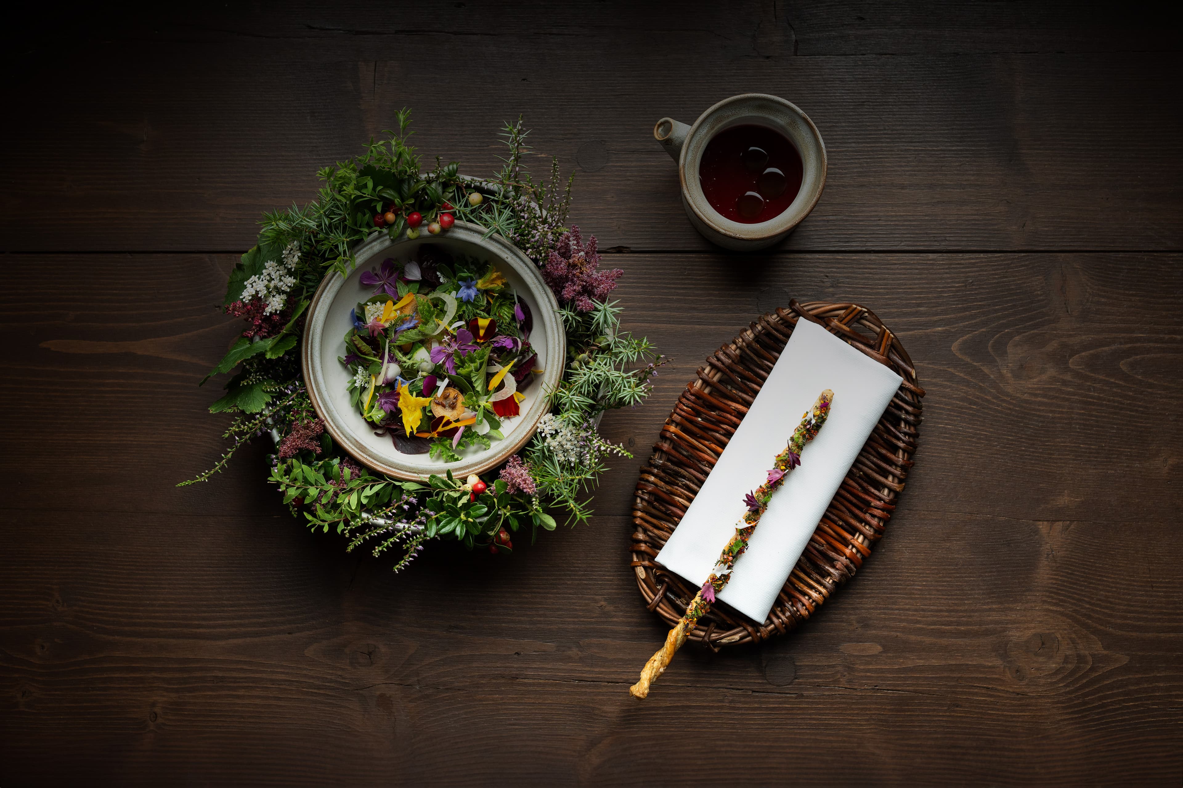 A round stone bowl of dressed wild herbs, mountain blossoms and edible flowers ringed in a wreath of foraged greenery on a dark oak table — beside it a wicker tray with a hand-rolled herb pastry, and a small ceramic cup of dark broth.