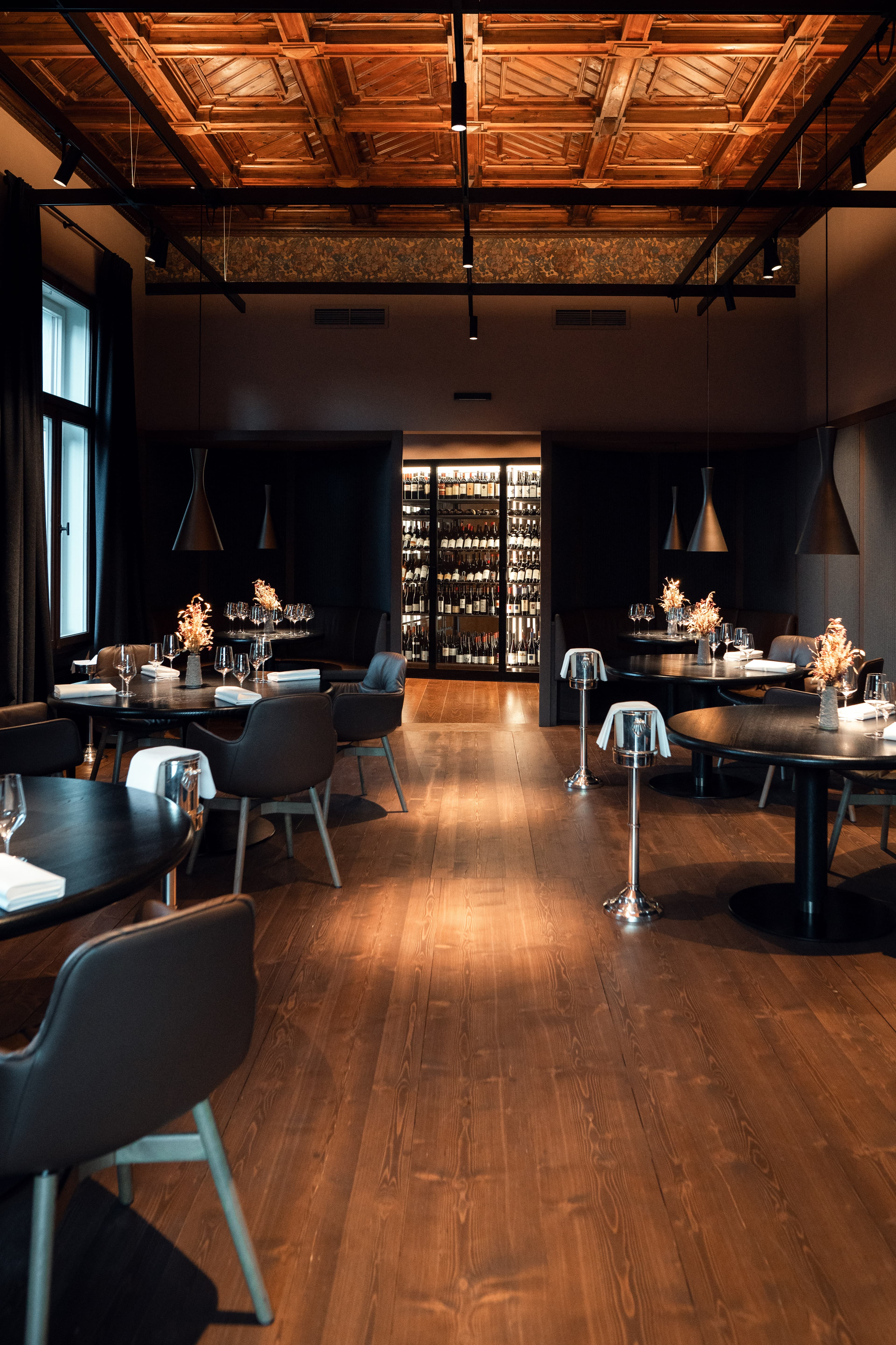The salon at Atelier Moessmer — high coffered wooden ceiling with a painted floral panel, two black pendant lamps, round black-topped tables on a polished plank floor, a lit wine wall at the far end of the room.