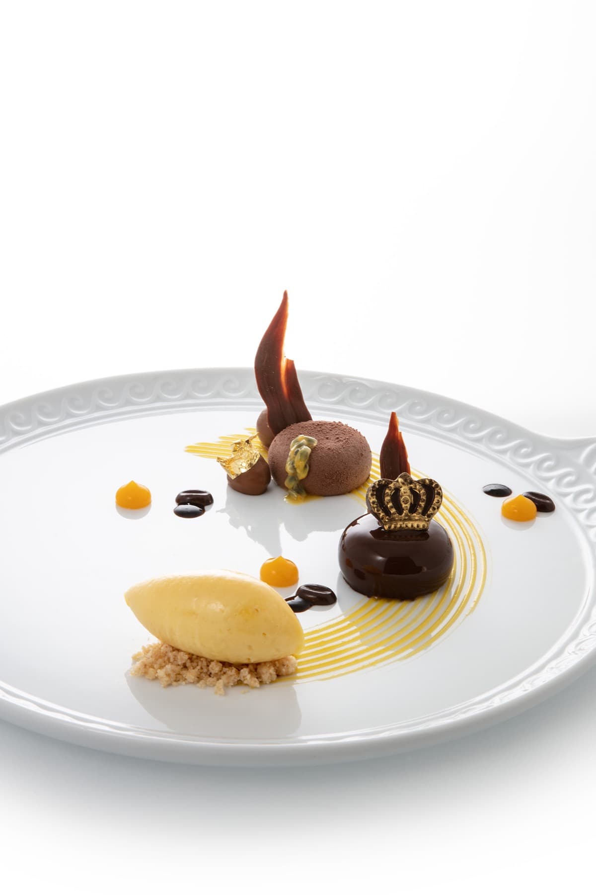 A chocolate dessert in the form of a small dome on a white scrollwork plate — a fine gold crown finial set on top, a sorbet quenelle on a hazelnut crumb, three drops of dark chocolate and a curve of saffron sauce around the plate.