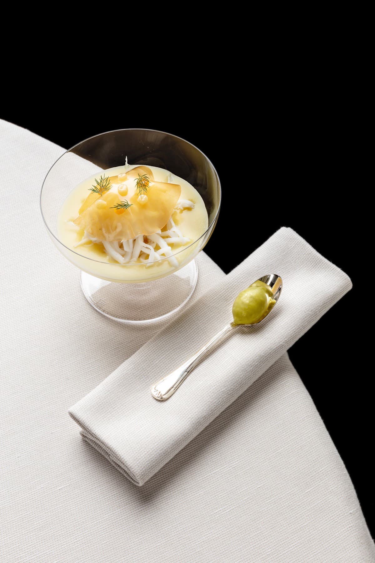 A small coupe glass on a white-linen napkin against black — a soft yellow custard at the base topped with shaved céleri and a thin slice of cured fish, dressed with dill, a silver spoon laid alongside with a dab of green oil.