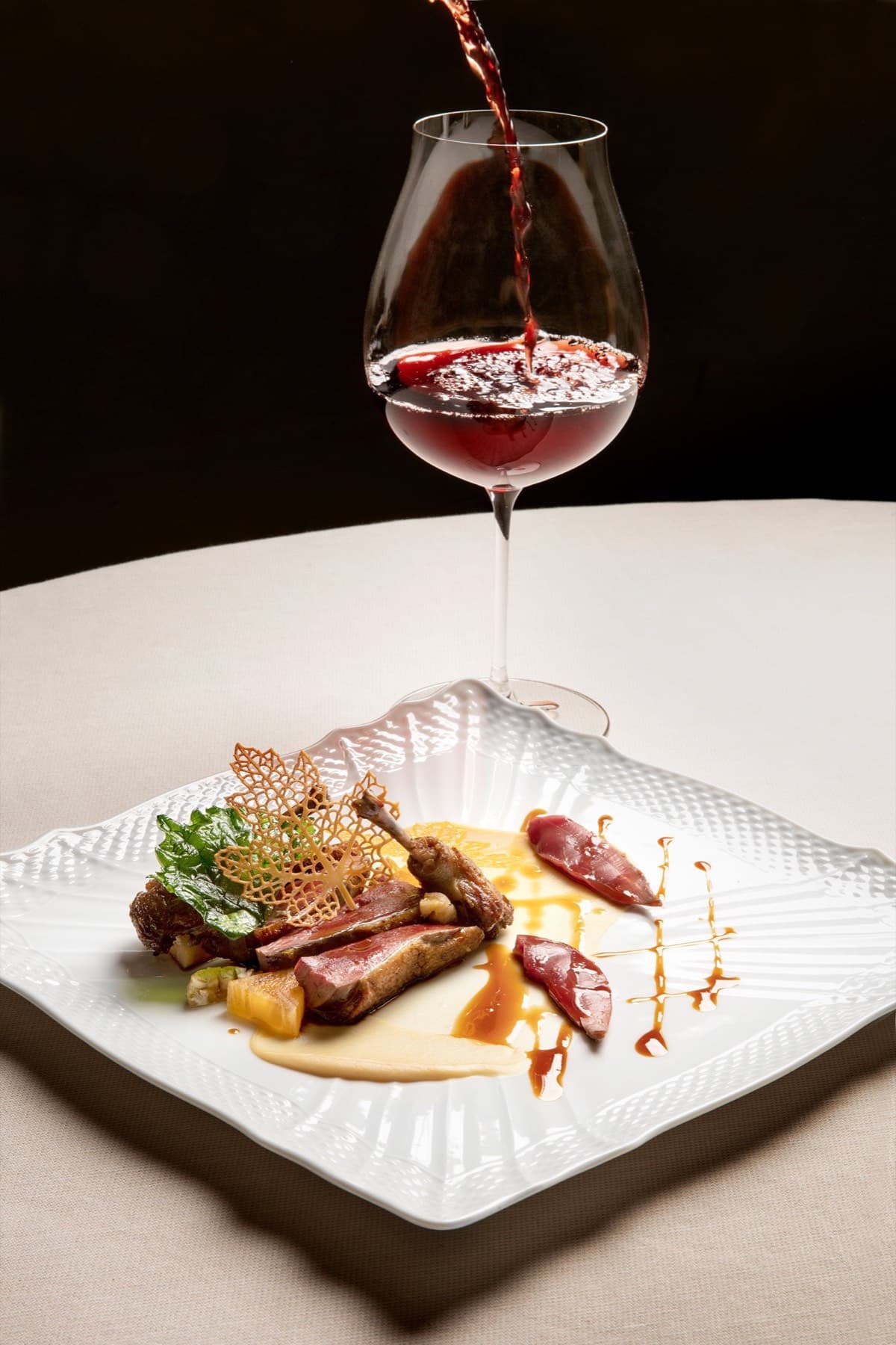 A scallop-rimmed plate of duck breast in three pink slices with caramelised pear, jus and a fine tuile of crisp lace — a glass of red Piemontese wine being poured behind the plate, the streaming wine catching the light.