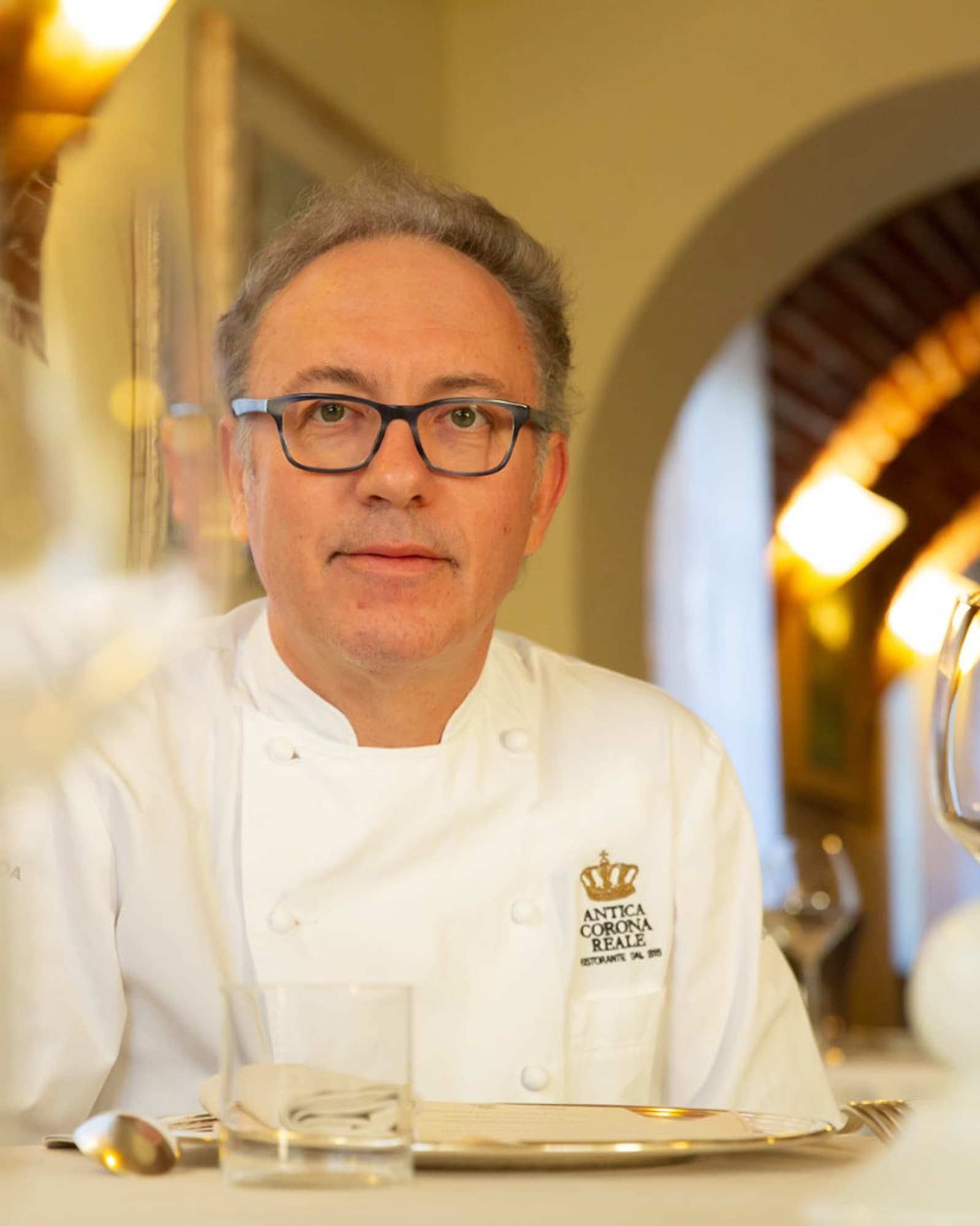 Portrait of chef Gian Piero Vivalda seated at a dining-room table in a white chef's jacket monogrammed Antica Corona Reale Ristorante dal 1815 — silver-grey hair, dark-rimmed glasses, a stone arch in warm light behind him.