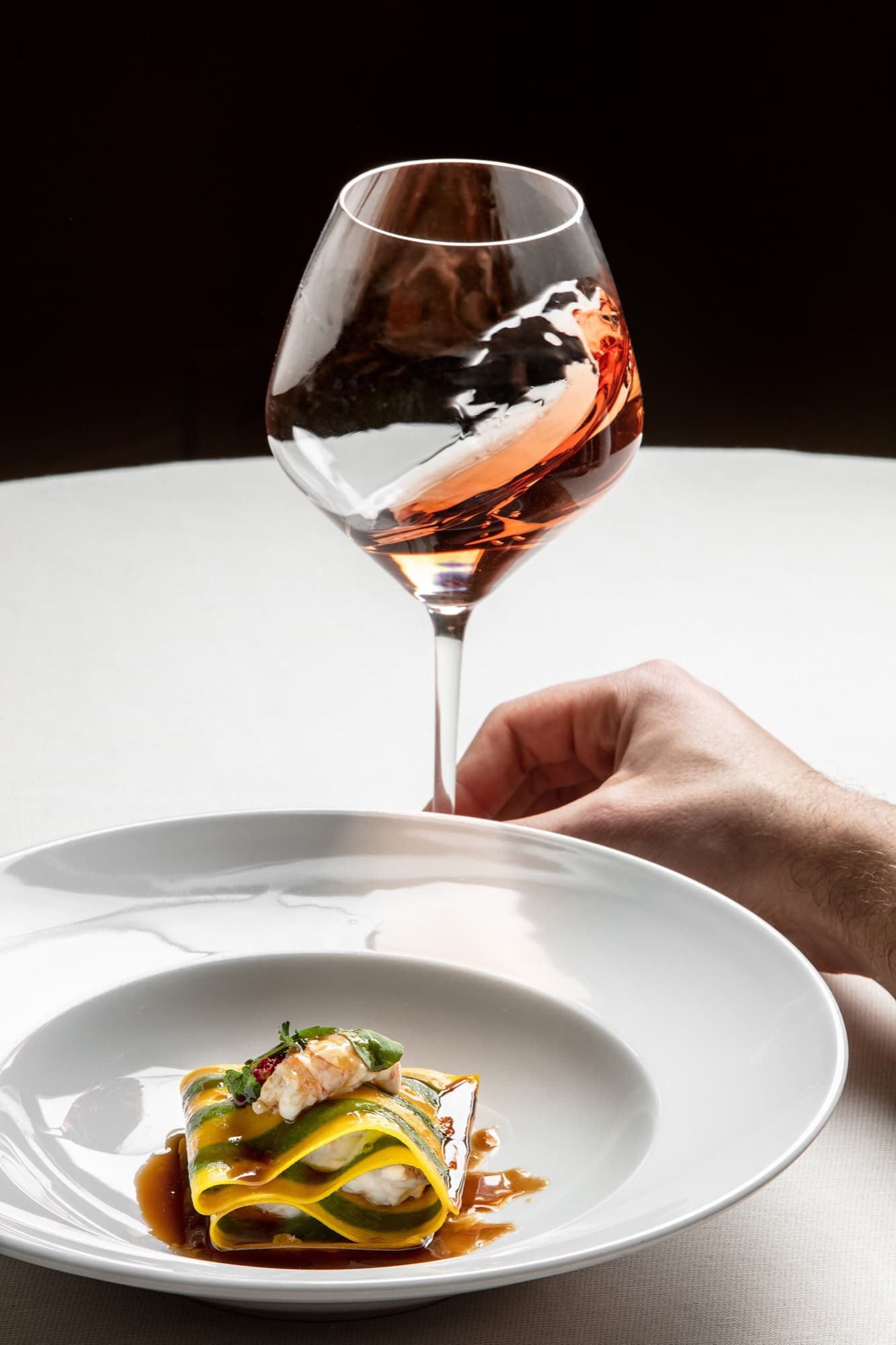 A pasta course at Antica Corona Reale — a small mille-feuille of yellow and green pasta sheets on a white plate, layered with prawn and dressed with a glossy bronze veal jus, a hand bringing in a large rosé wine glass above it against a dark background.