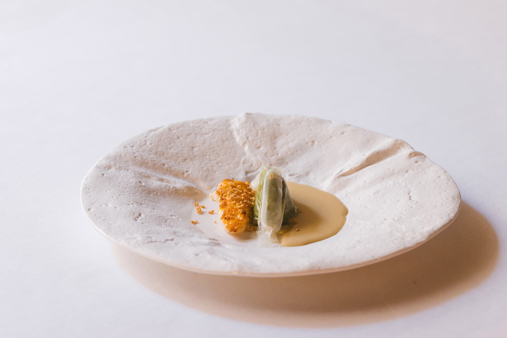 A signature plate at La Madia — a textured cream-coloured ceramic dish, the surface like crumpled paper, set with a small piece of crisped fish, a lettuce roll and a pool of pale sauce, photographed against a white linen cloth.