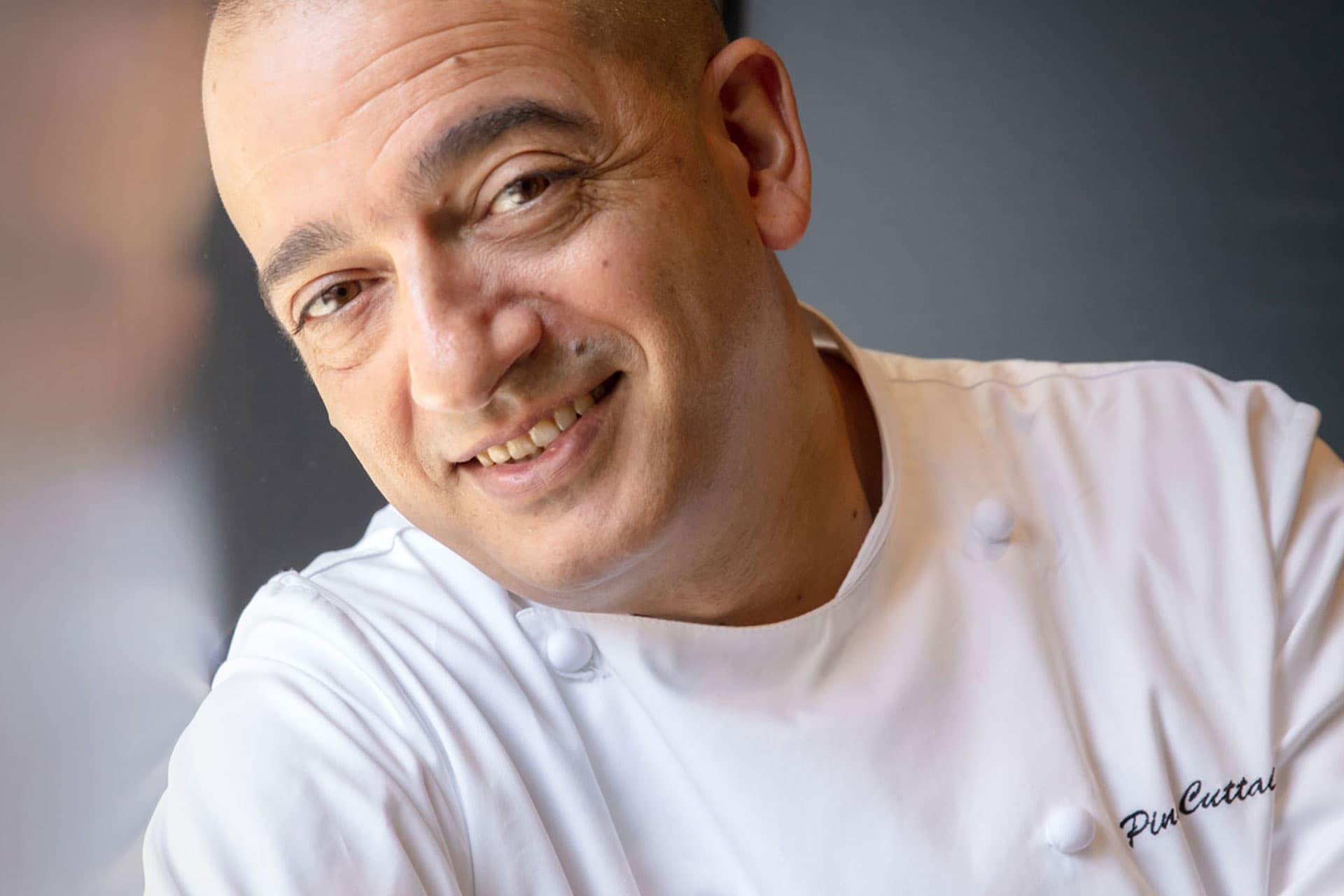 Chef Pino Cuttaia in close portrait — head shaved, broad smile, in chef's whites embroidered Pino Cuttaia, photographed against a soft grey wall.